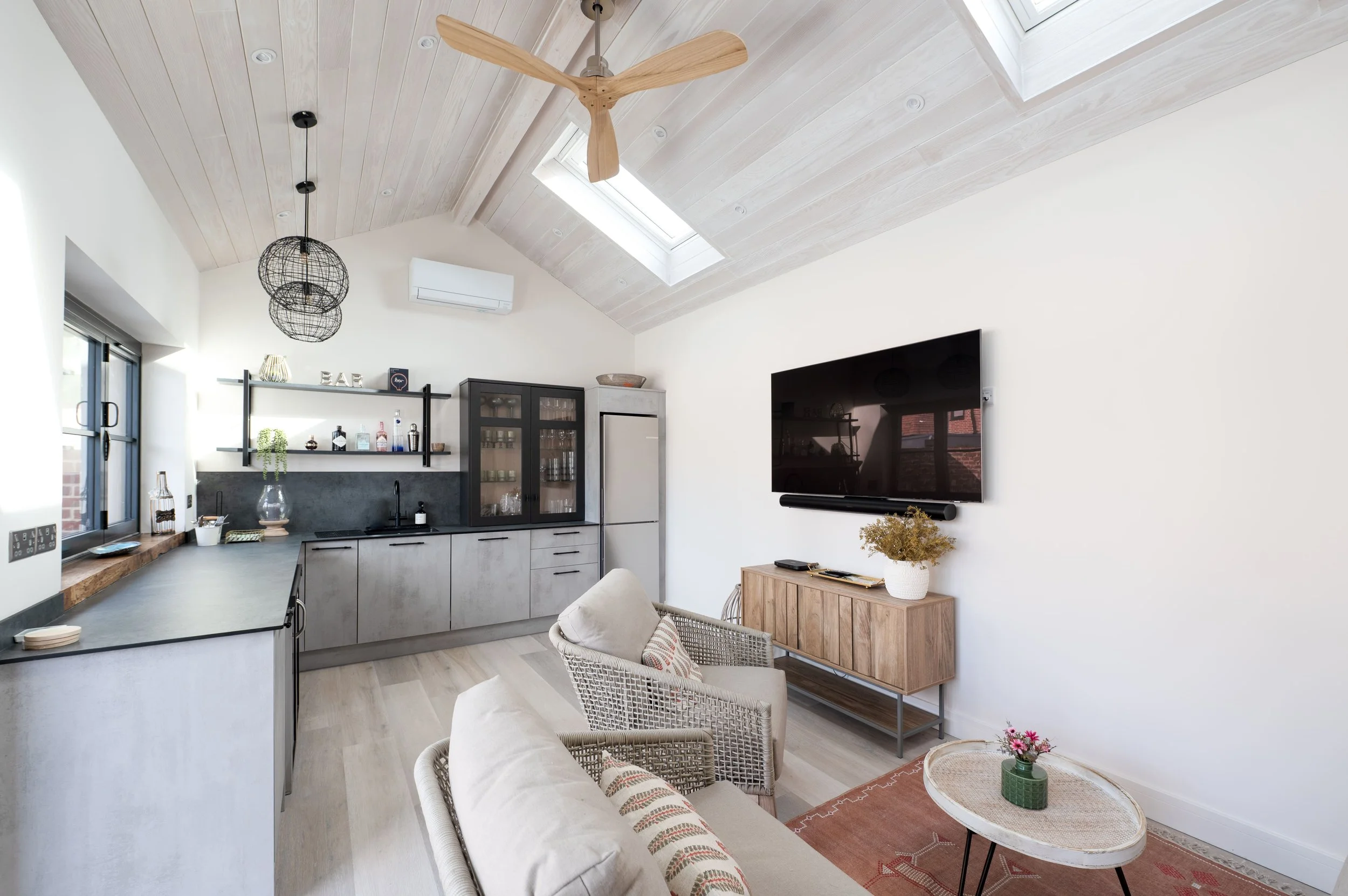 Living room with a mounted flat-screen TV, a wooden console table, two armchairs with cushions, a small round coffee table with pink flowers, a kitchen area with a black countertop, shelves with bottles and decor, a refrigerator, a ceiling fan, skylights, and hanging light fixtures.