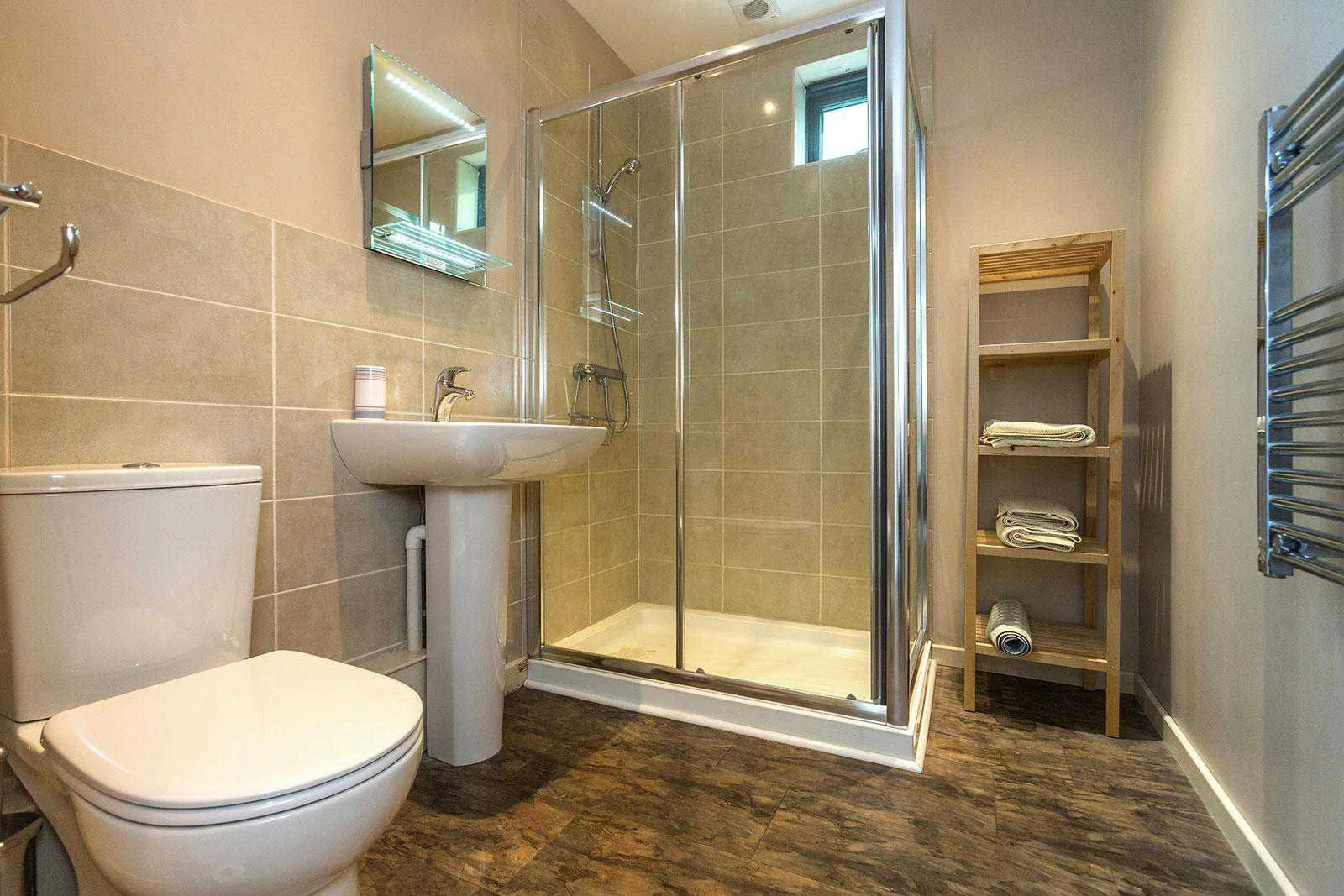 A modern bathroom with beige tiled walls, a white toilet, and a white pedestal sink. There is a shower with a glass door, a small window above the shower, and a wooden shelving unit with folded towels and a rolled towel. The flooring is dark wood laminate.