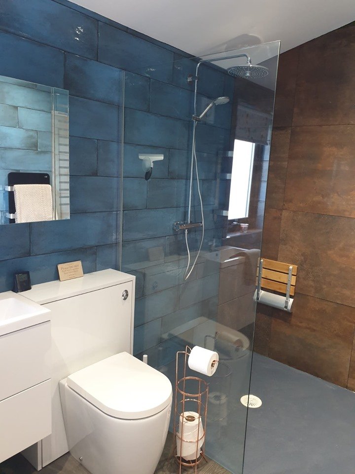 Modern bathroom with a blue tiled wall, a white toilet, a small mirror, and a shower area with a rainfall showerhead and handheld shower. There is a wooden bench and a glass partition separating the shower from the rest of the bathroom. A toilet paper holder with spare rolls is nearby.