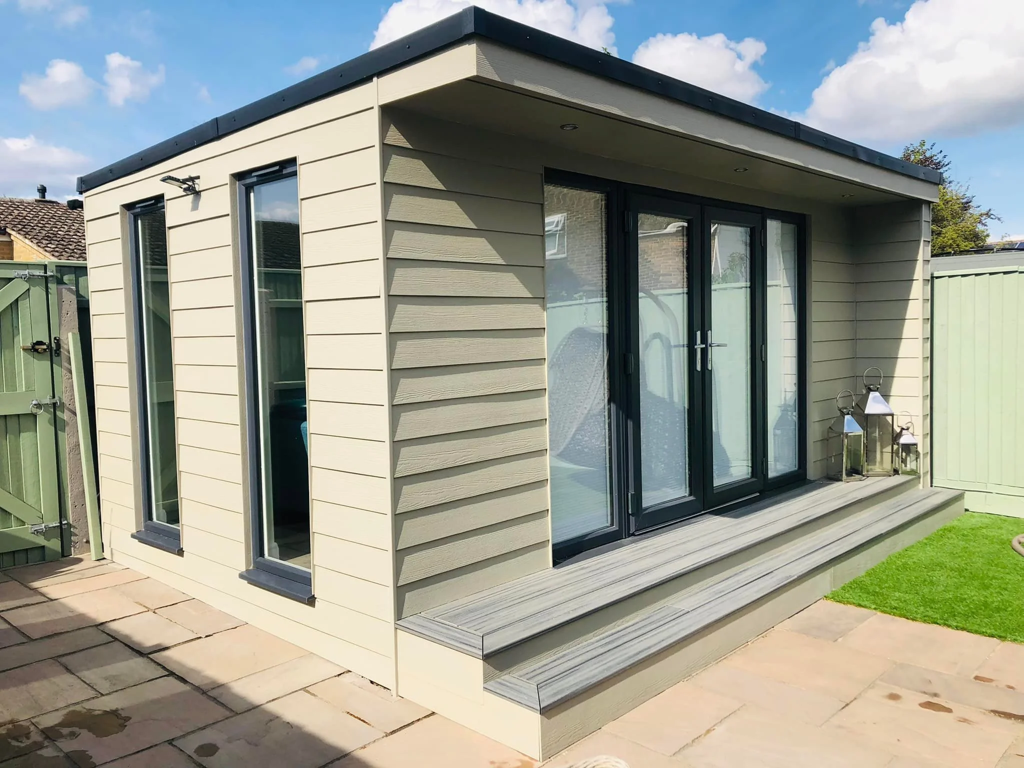 A modern small garden building with beige horizontal siding and large glass sliding doors, situated on a wooden deck with steps, surrounded by a paved patio, green lawn, and a light green fence, under a blue sky with clouds.