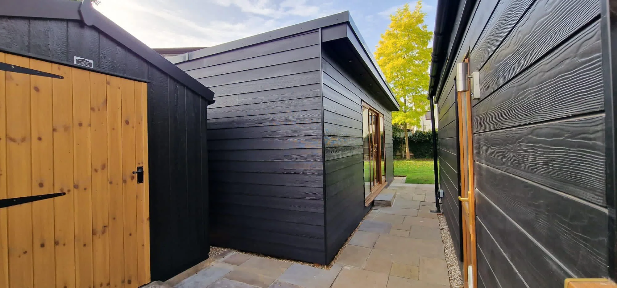 View of a modern backyard with black and natural wood siding outbuildings and a gray paved walkway, with a green tree and blue sky in the background.