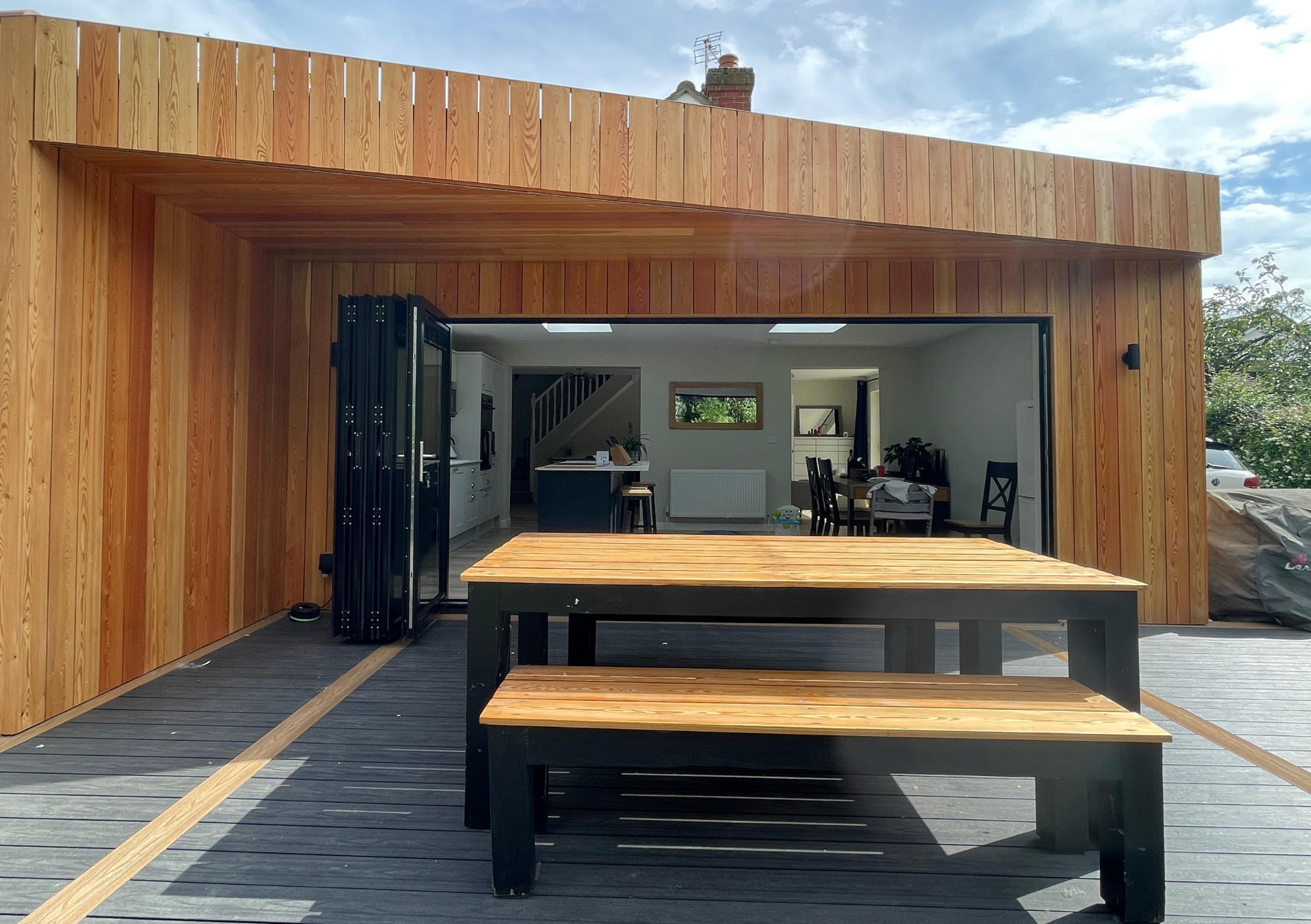 View of a modern wooden patio leading to an open indoor living space with a dining table, chairs, and a kitchen area.