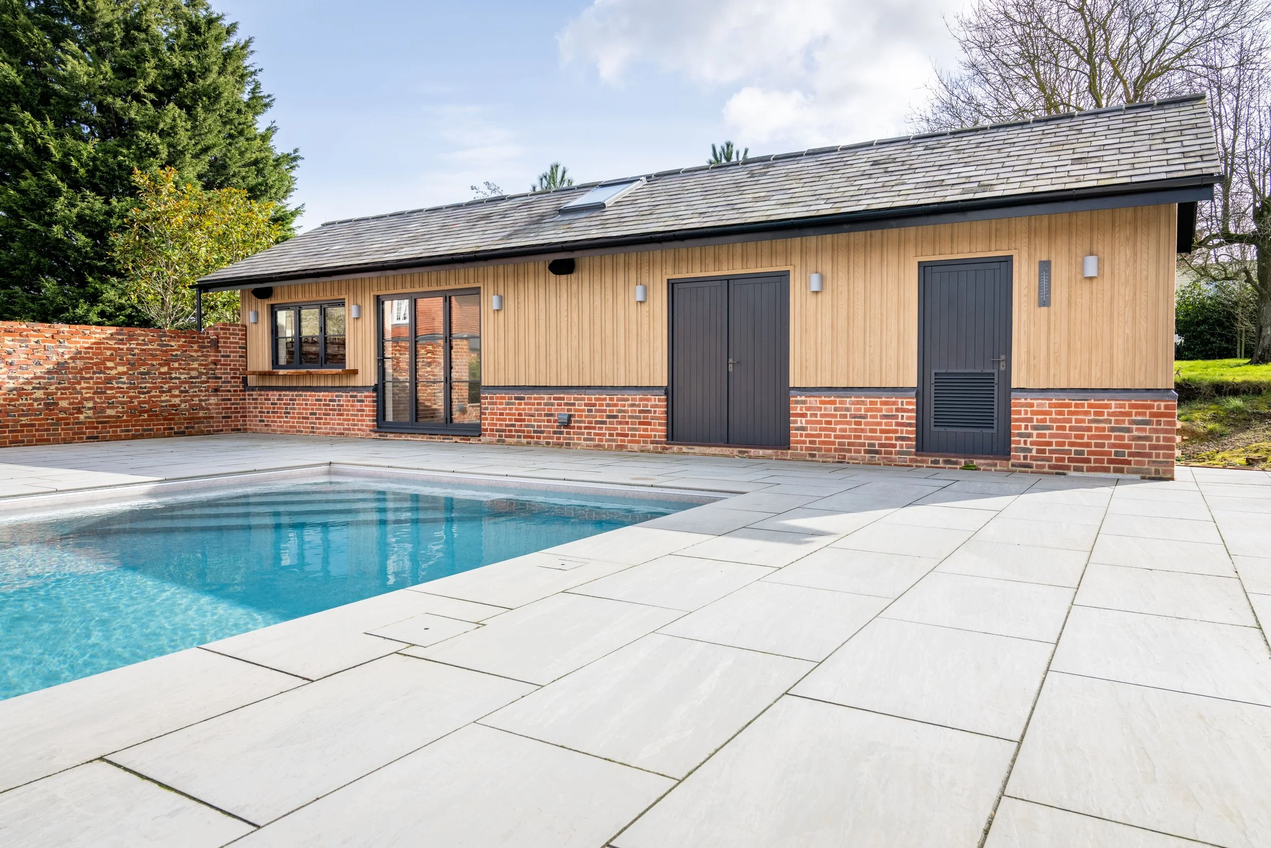 Modern backyard with a swimming pool, patio, and a house featuring wood siding, brick accents, and black doors.