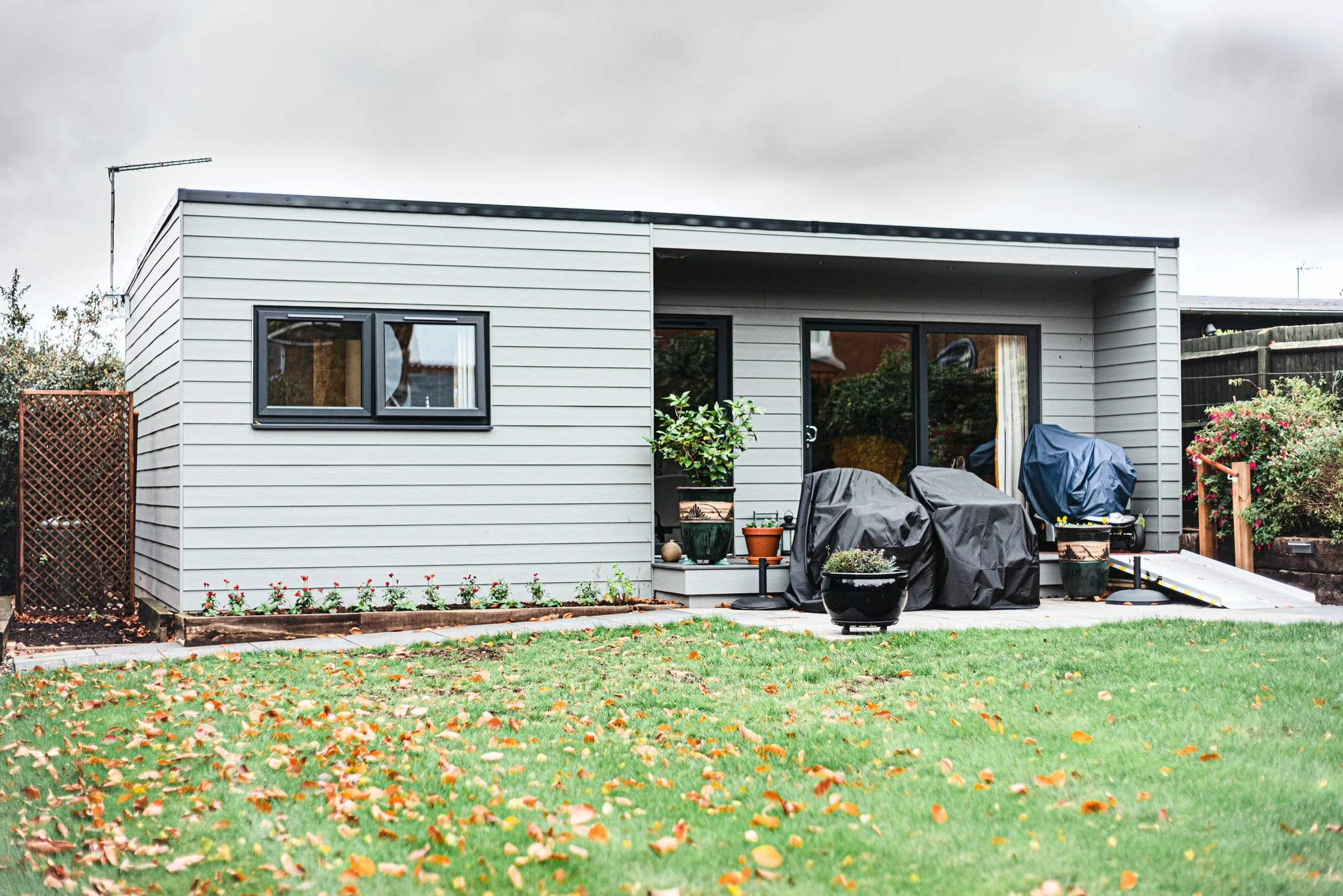 A small modern house with a gray exterior, sliding glass doors, and a small backyard with a grass lawn and fallen leaves. There are two covered barbecue grills, potted plants, and a ramp leading to the back door.