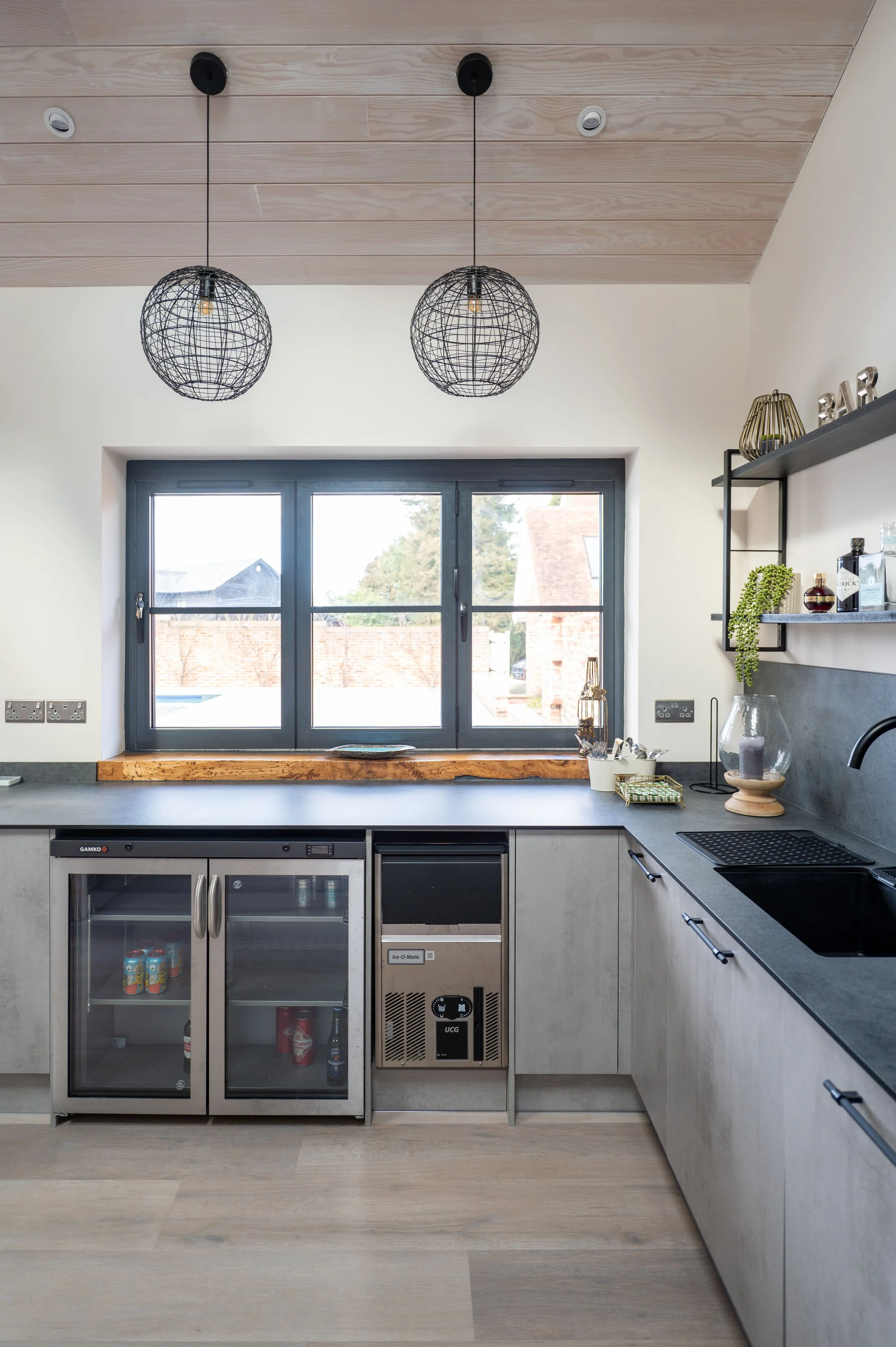 Modern kitchen with black-framed window, concrete countertops, and minimalist decor. Two black wire pendant lights hang from a wood-paneled ceiling.