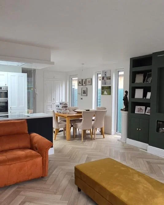 Living and dining area with orange sofa, yellow ottoman, dining table with cream chairs, dark green built-in shelves, and framed artwork on the walls.