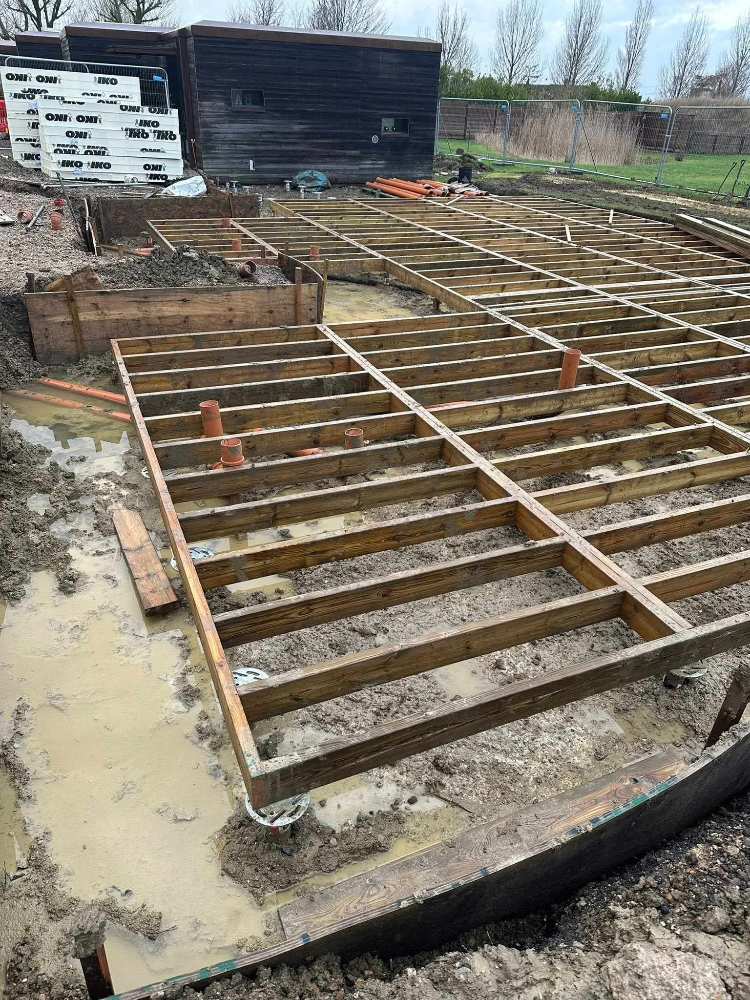 Construction site with wooden foundation framing for a building, with muddy and wet ground, some orange pipes, and a black shed in the background.