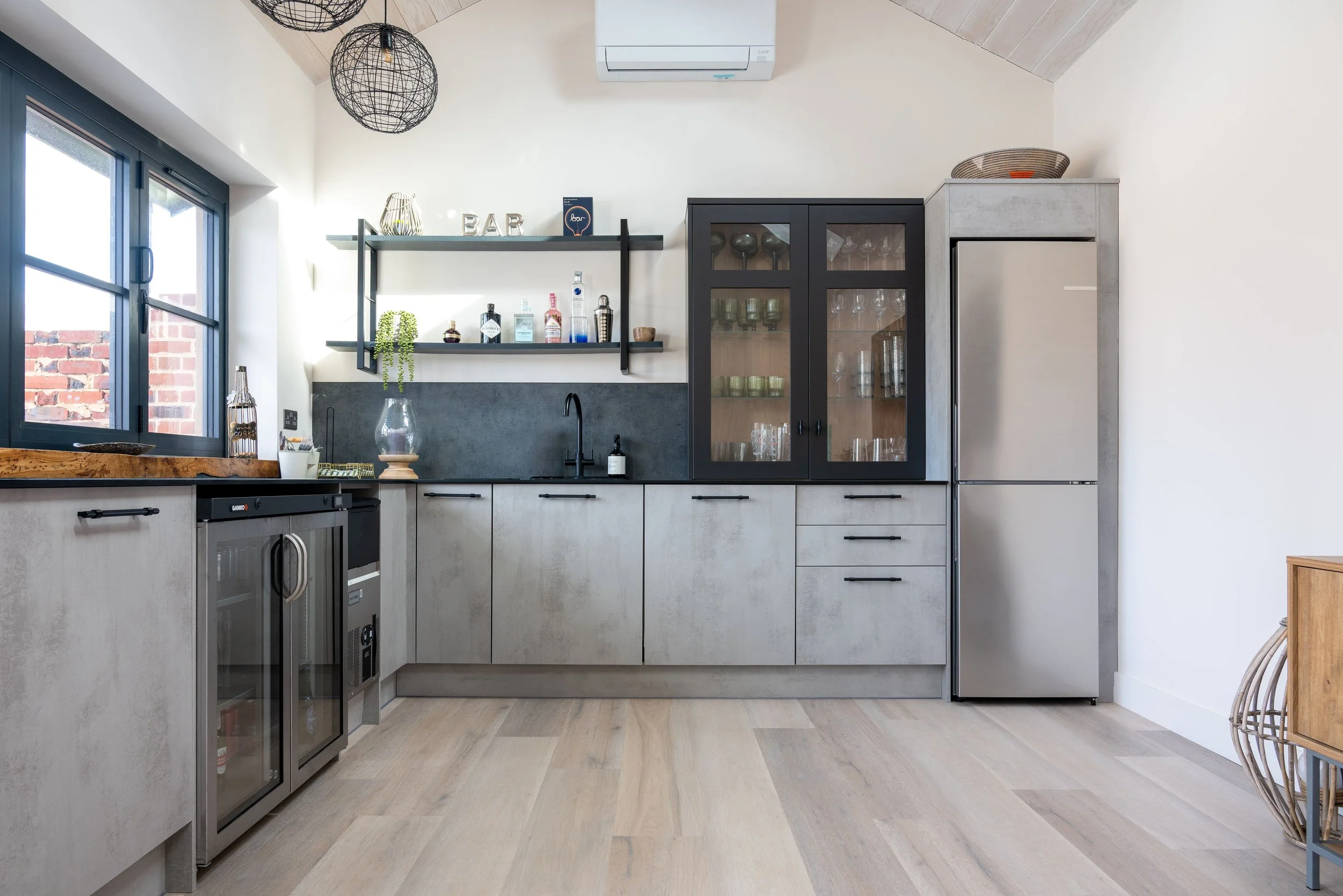 Modern kitchen with black and concrete cabinetry, open shelving with decor, black framed window, black pendant lights, wooden countertop, stainless steel appliances, and light wood floor.