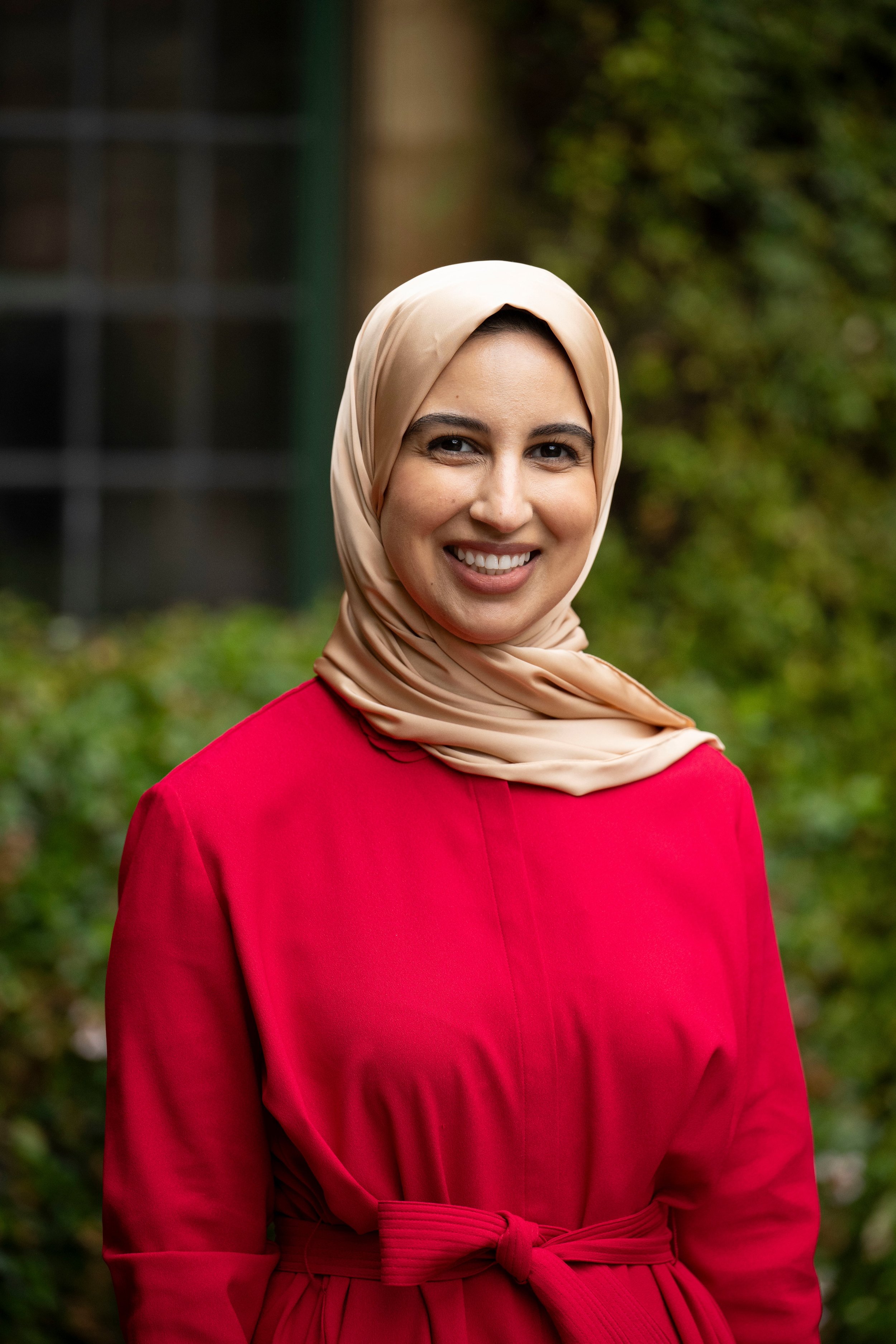 A woman wearing a beige hijab and a bright red dress, smiling outdoors with greenery in the background.