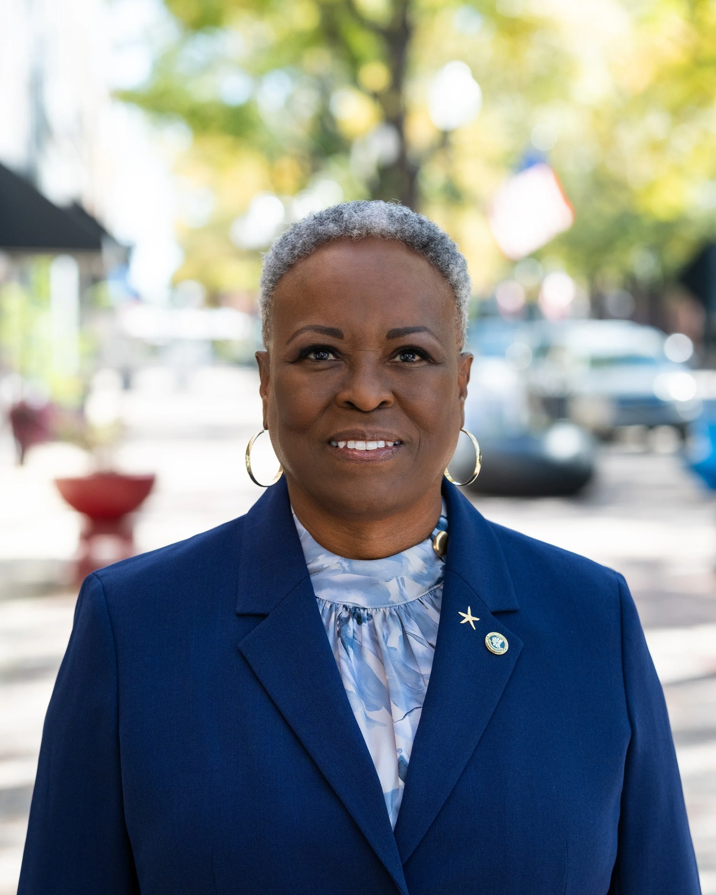A woman with short gray hair wearing a dark blue suit and a patterned blouse, standing outdoors with trees and parked cars in the background.