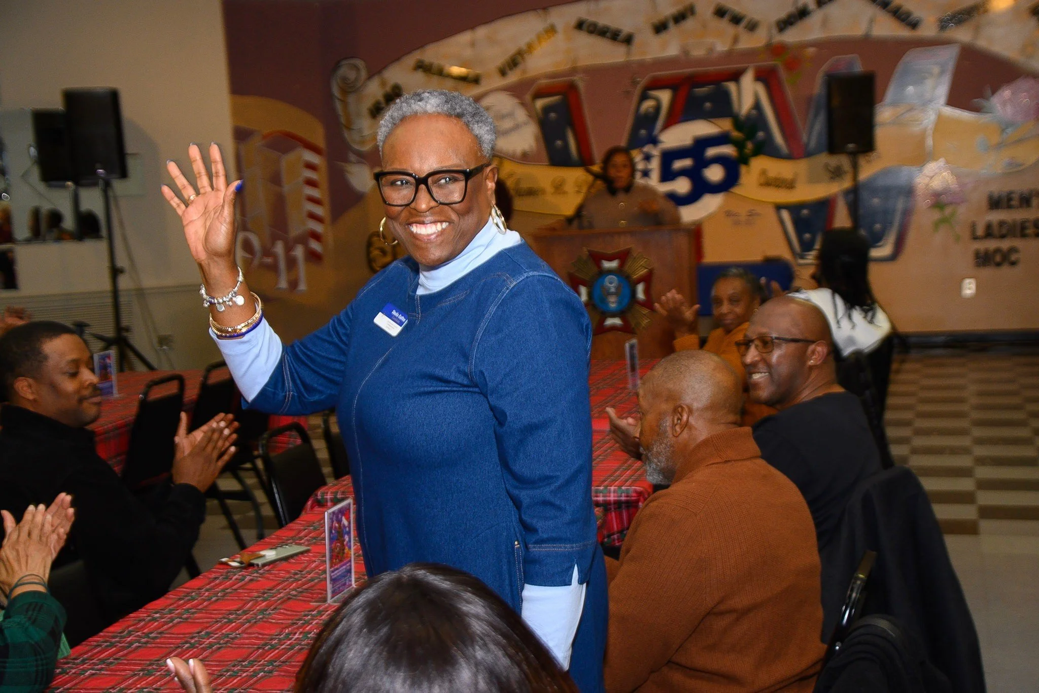 A woman with gray hair, glasses, and earrings wearing a blue jacket is smiling and waving at the camera, sitting at a table with other people in a decorated room with a mural in the background.