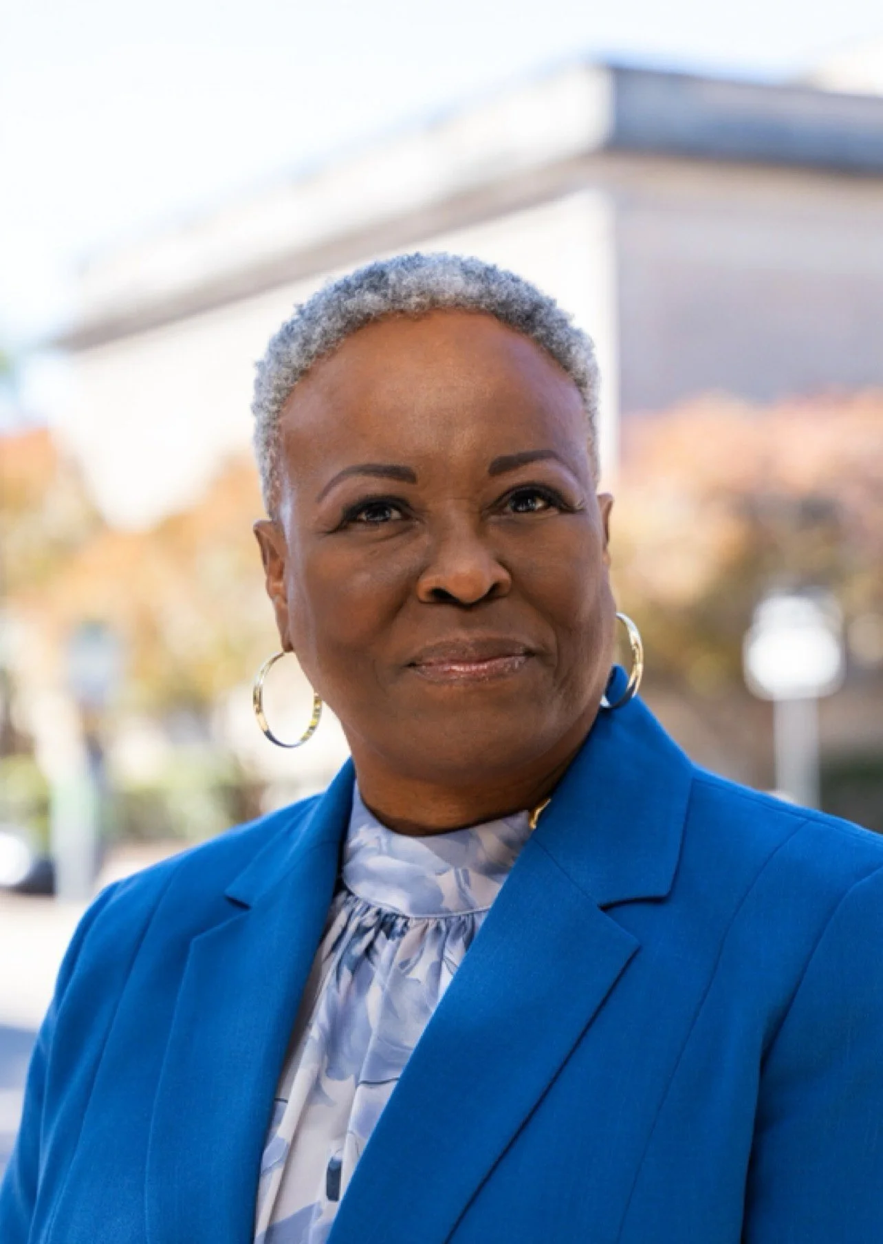 Close-up of a confident middle-aged African American woman with short gray hair, wearing a blue blazer, hoop earrings, and a patterned blouse, standing outdoors on a sunny day with blurred trees and building in the background.