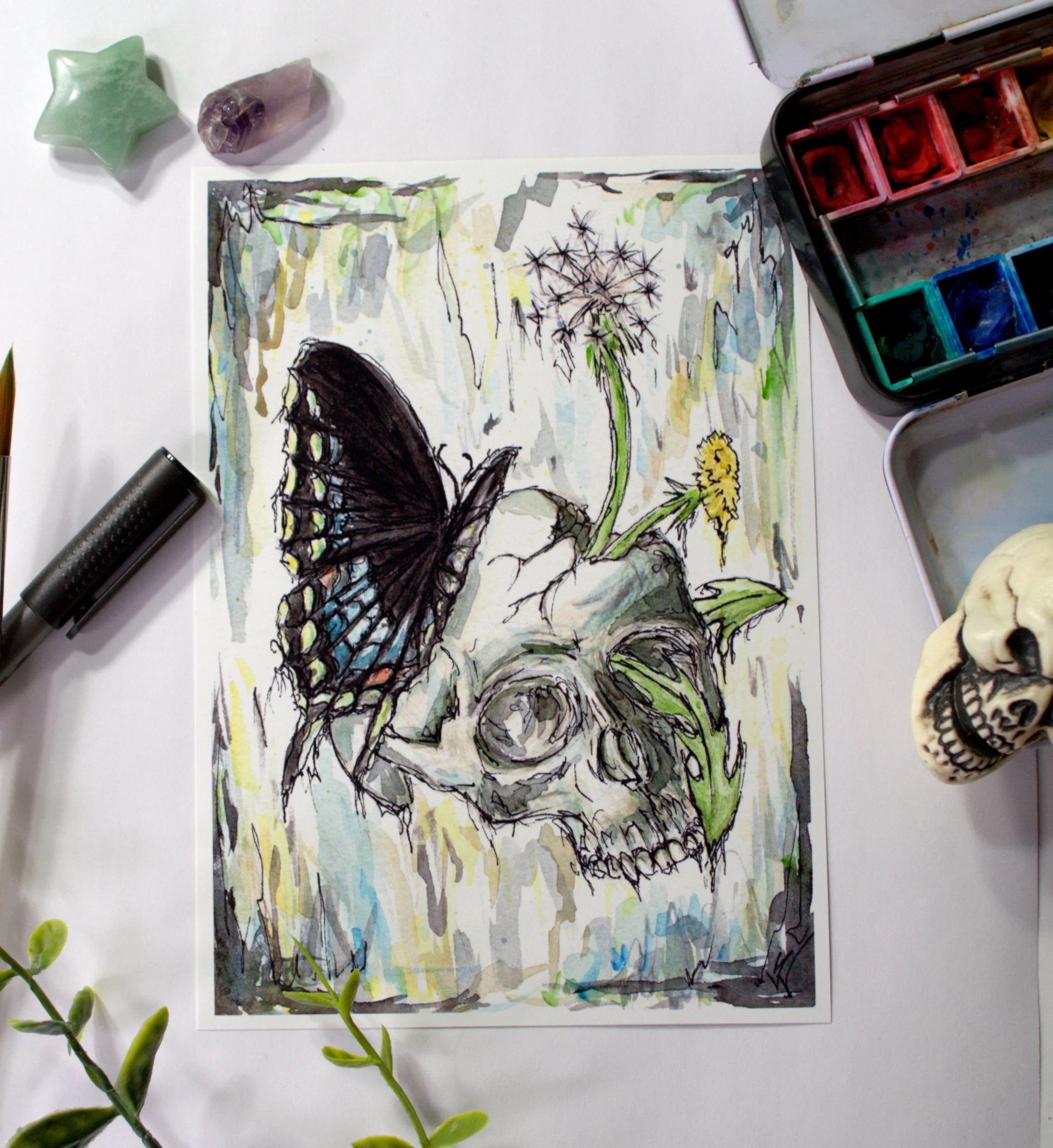 A watercolor painting of a black butterfly perched on a forgotten skull with a dandelion flower growing through a crack.