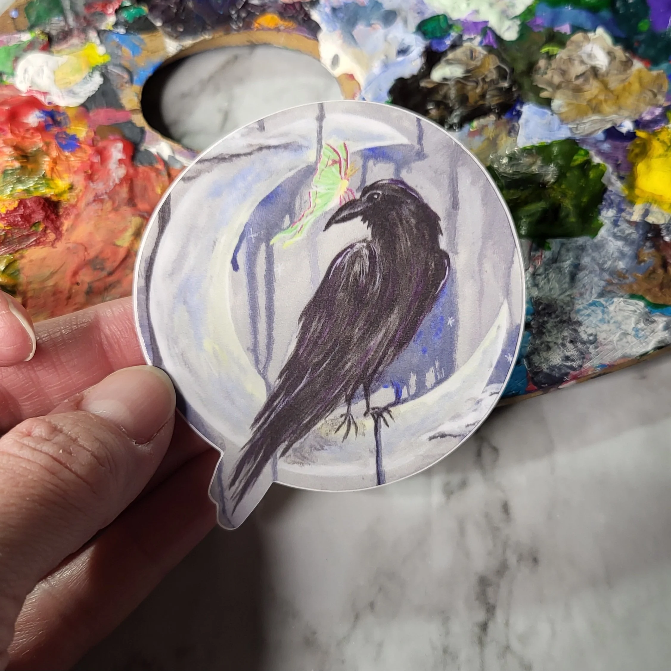 A person holding a circular pin with an illustration of a black bird, possibly a raven, standing on the ground with its beak open, in front of a colorful, textured background with paint smudges and streaks.