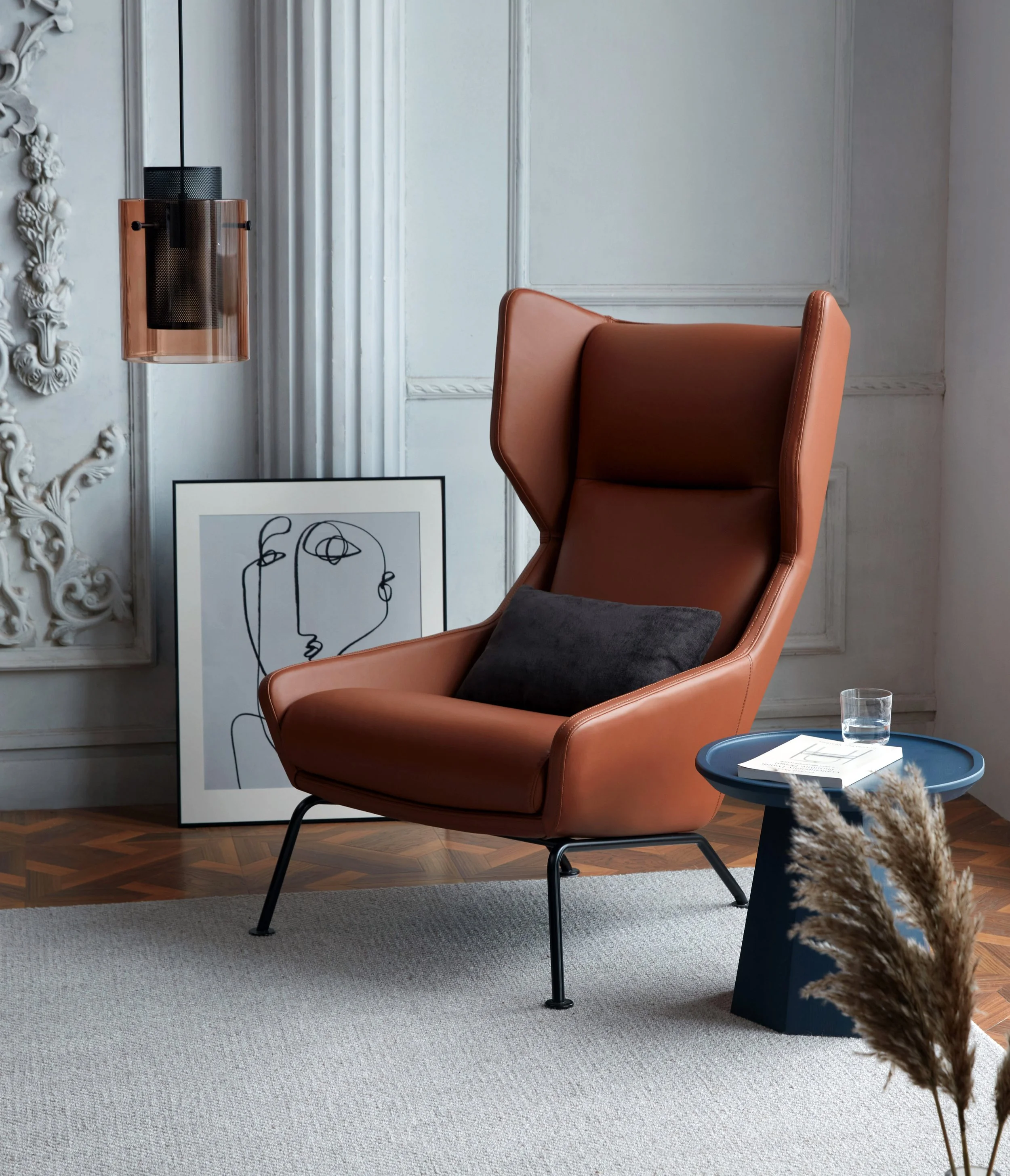 A modern interior with a high-back leather armchair, a black sofa cushion, a blue side table with a glass of water and papers, a framed abstract line art drawing of faces leaning against the wall, decorative wall molding, and a pendant light with a c