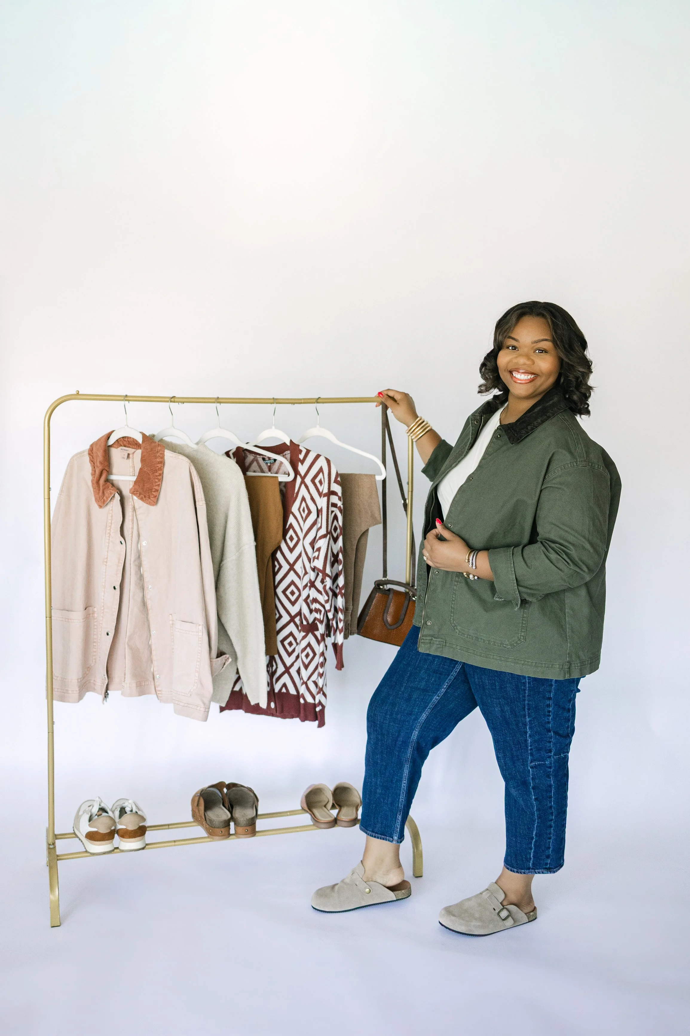Woman smiling next to a clothing rack with jackets, sweaters, and shoes.