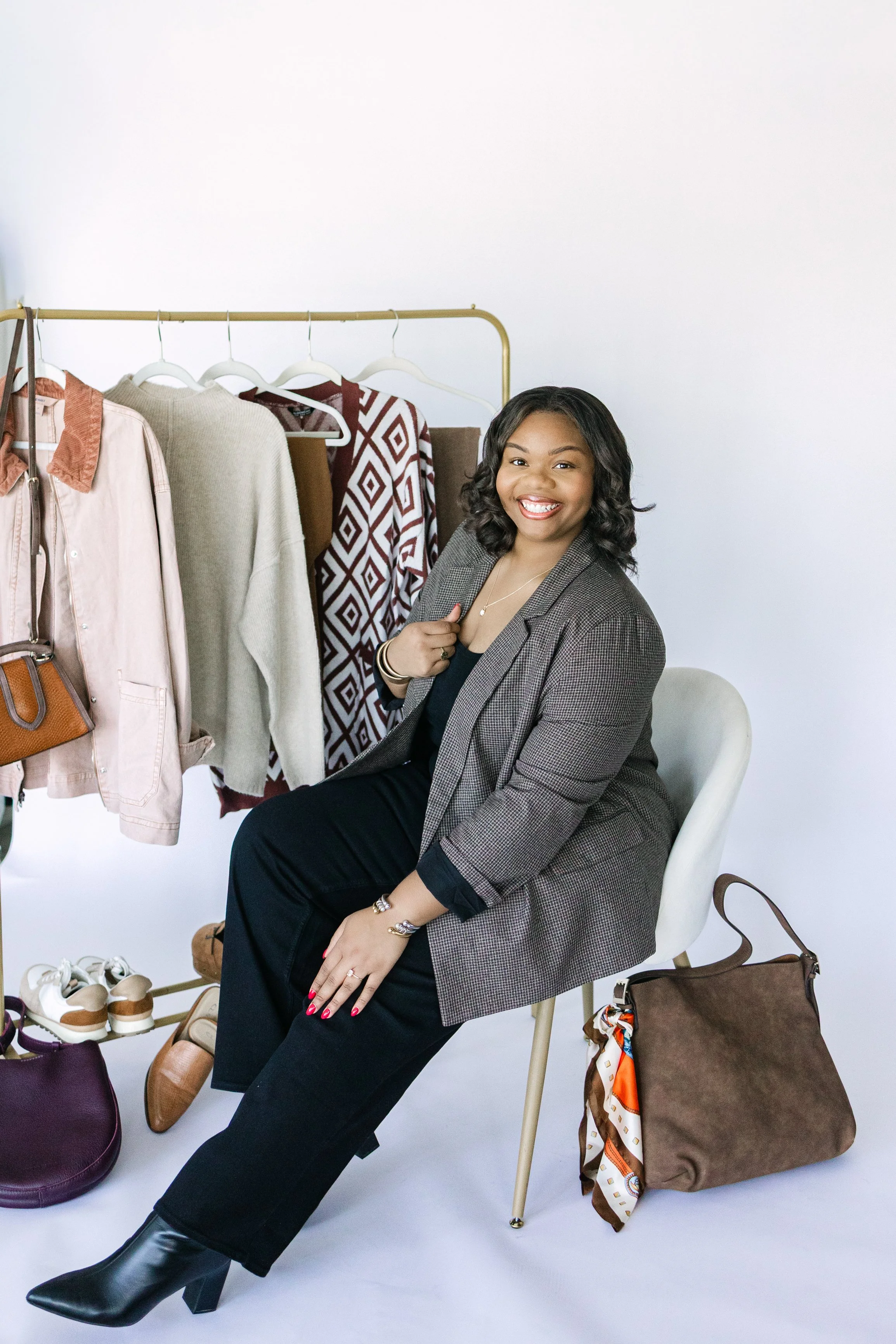A woman sitting on a white chair in front of a clothing rack with various garments, smiling at the camera, with bags on the floor.