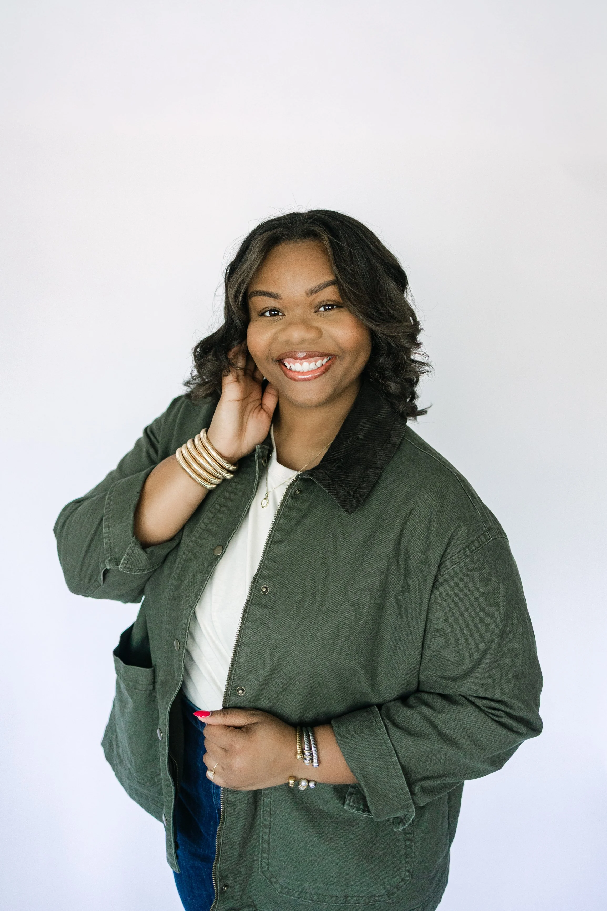 A smiling woman with wavy black hair wearing a green jacket with a black collar, a white shirt, and gold jewelry, standing against a plain white background.