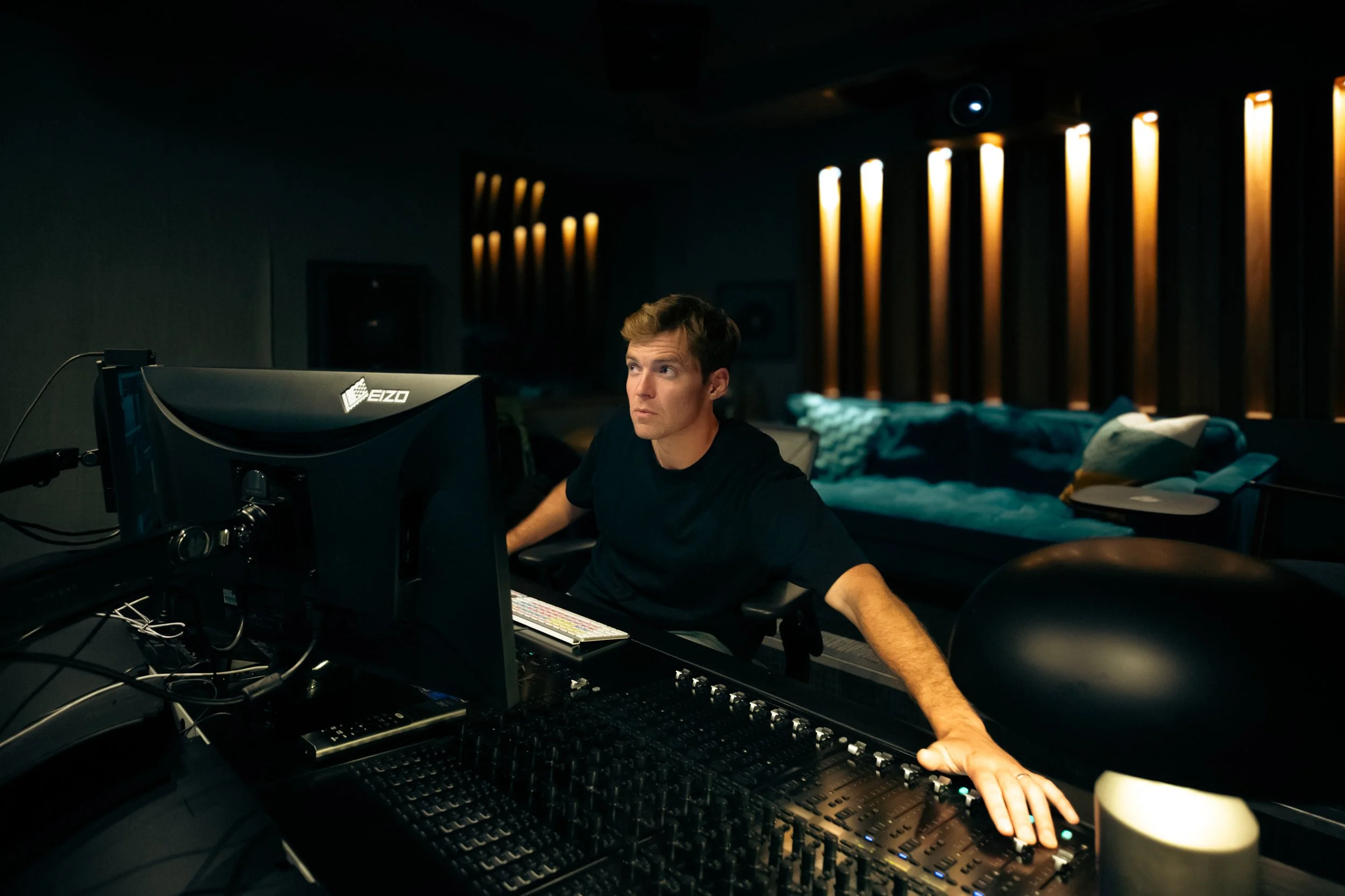 A man working at a sound mixing console in a dimly lit studio with a couch in the background.