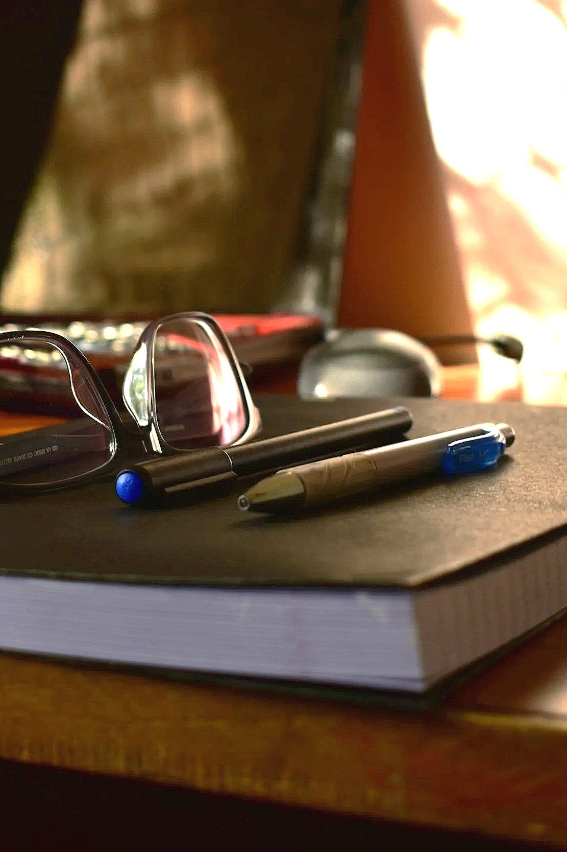 A pair of reading glasses, a blue pen, and a mechanical pencil resting on a closed black notebook on a wooden surface, with a computer screen and a speaker in the background.