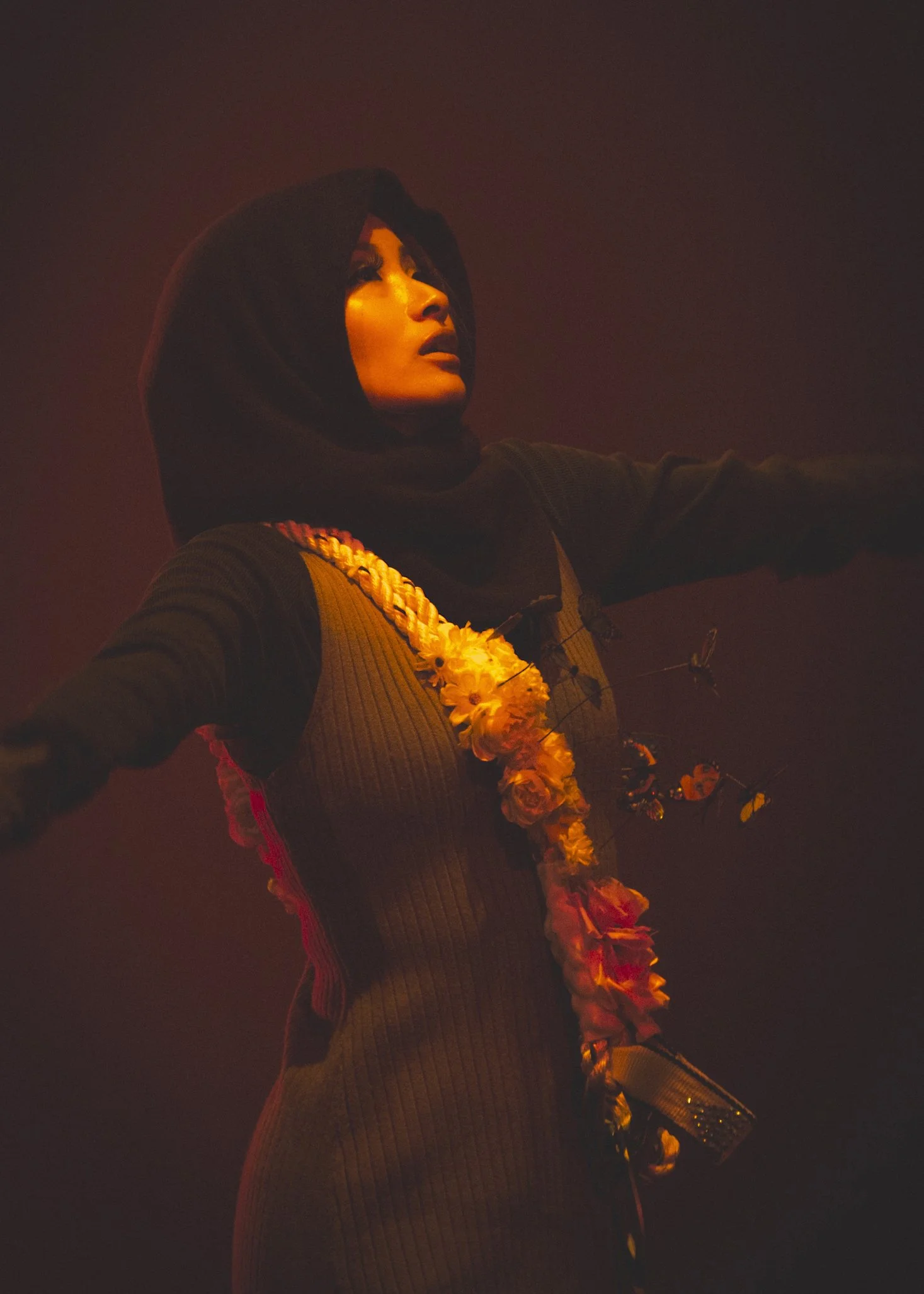 A woman in a black headscarf and long-sleeved dress, wearing a floral garland around her neck, standing with arms outstretched in a dimly lit, smoky environment.