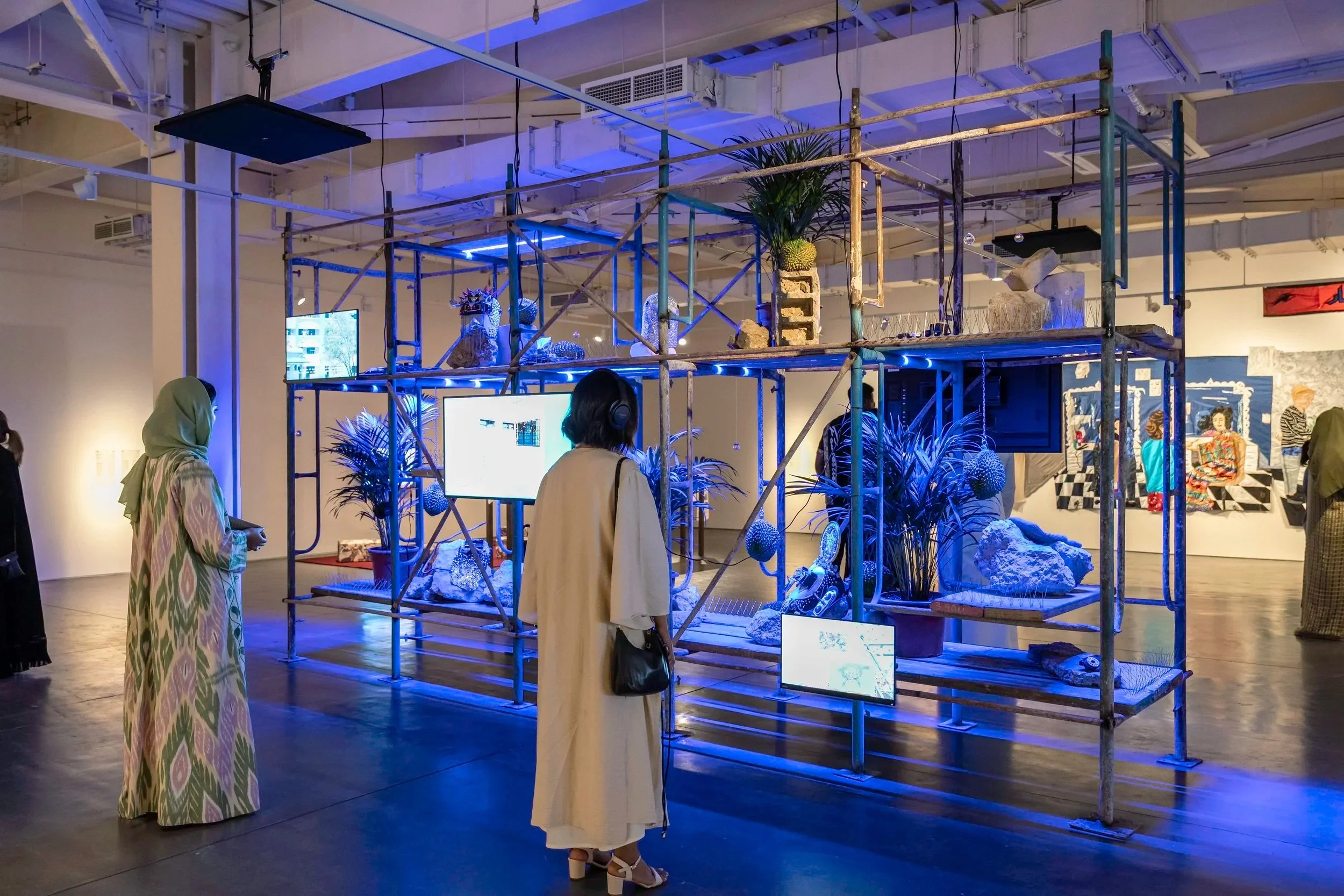 Art installation in a gallery with scaffolding, blue lighting, sculptures, plants, and digital screens, with visitors observing.
