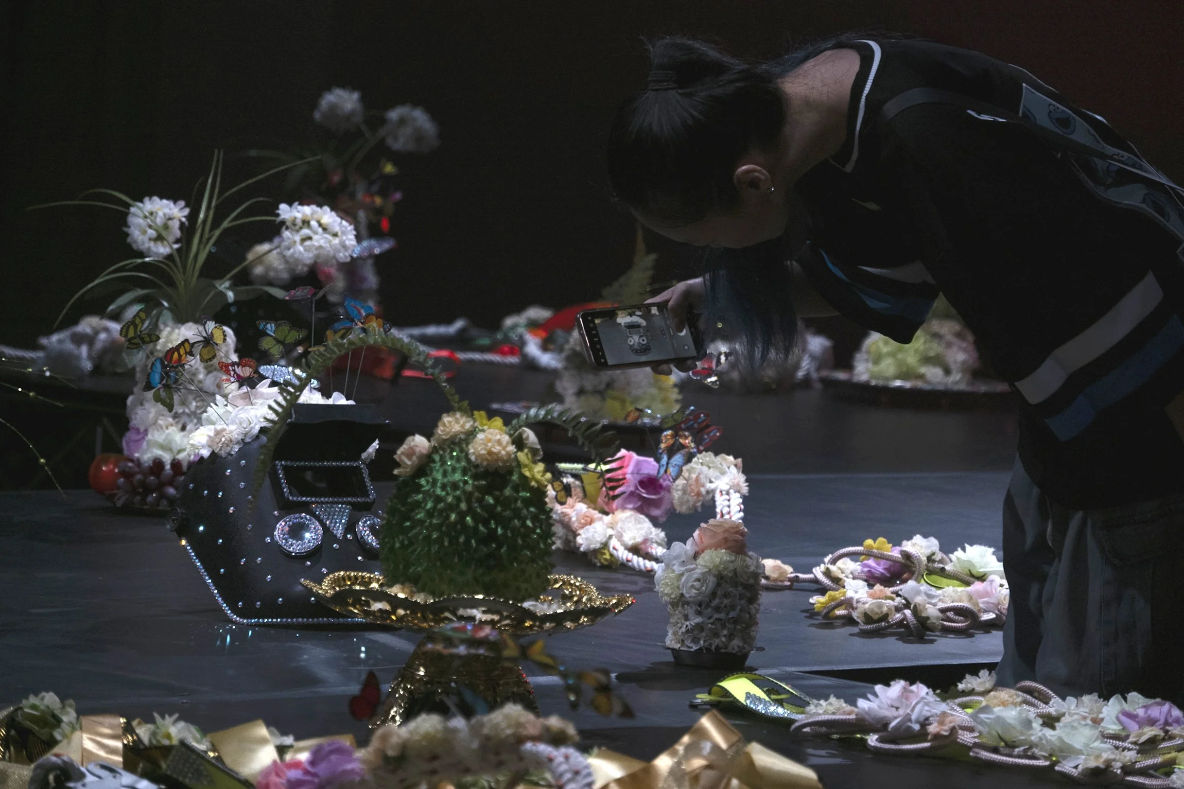 A woman is taking a photo of floral arrangements and jewelry on a black table with her phone. The arrangements include flowers, jewelry, and decorative butterfly ornaments.