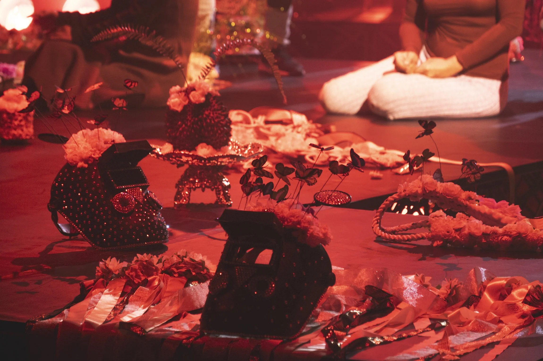 Decorative shoes and accessories, including miniature car-shaped shoes with floral embellishments, displayed on a table with people kneeling in the background, under warm lighting.