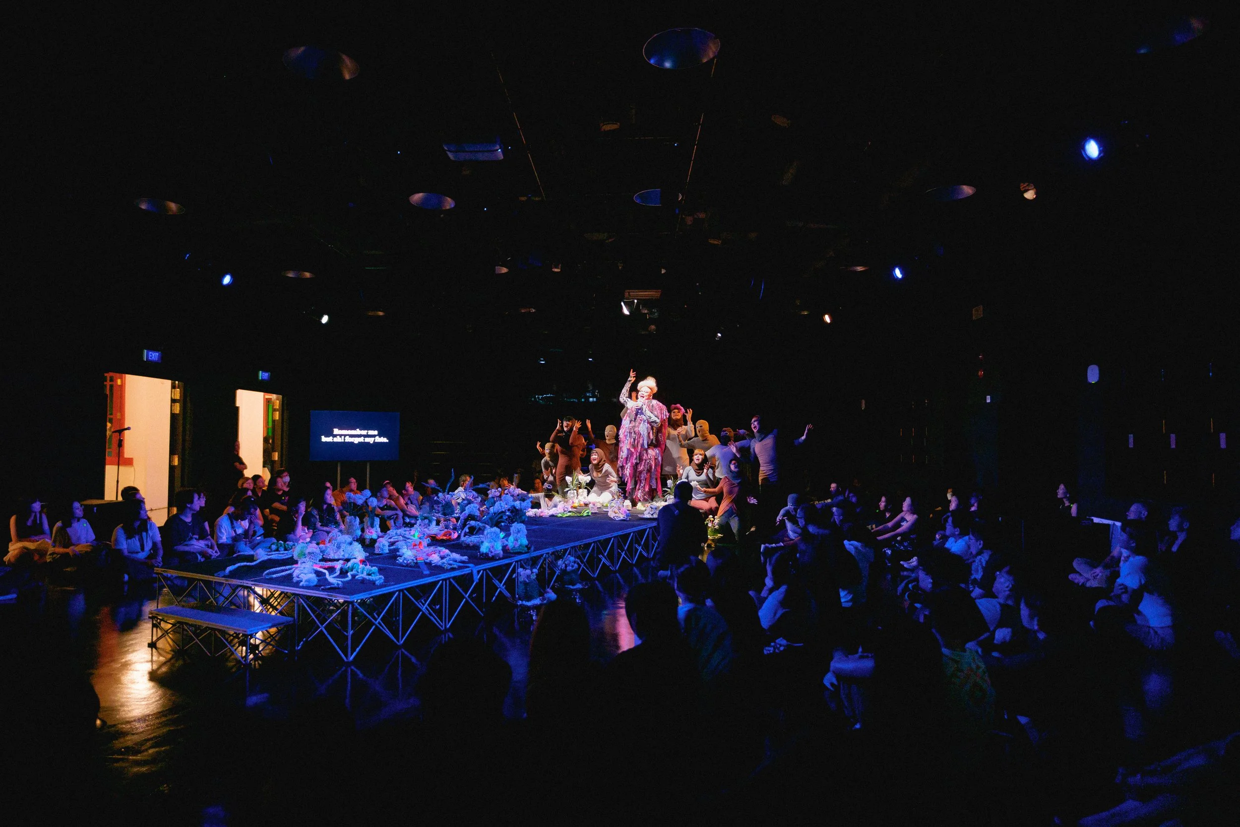 Theater stage with performers in colorful costumes surrounded by audience in a dark room illuminated by dim lighting.