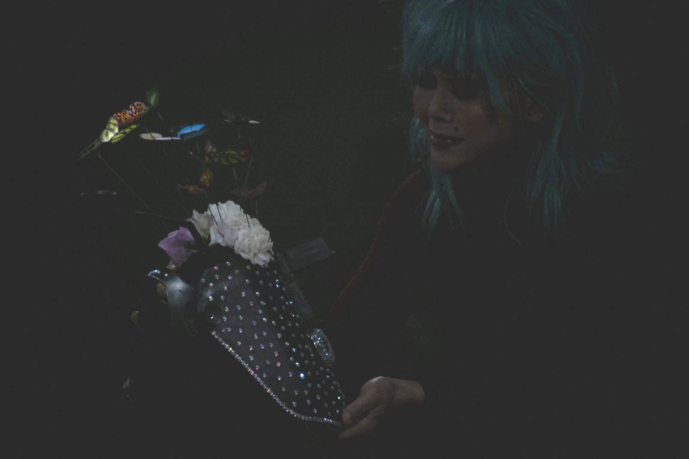 A person with bright blue hair and dark clothing sitting in a dark room, with a decorated bicycle that has flowers, butterflies, and rhinestones.