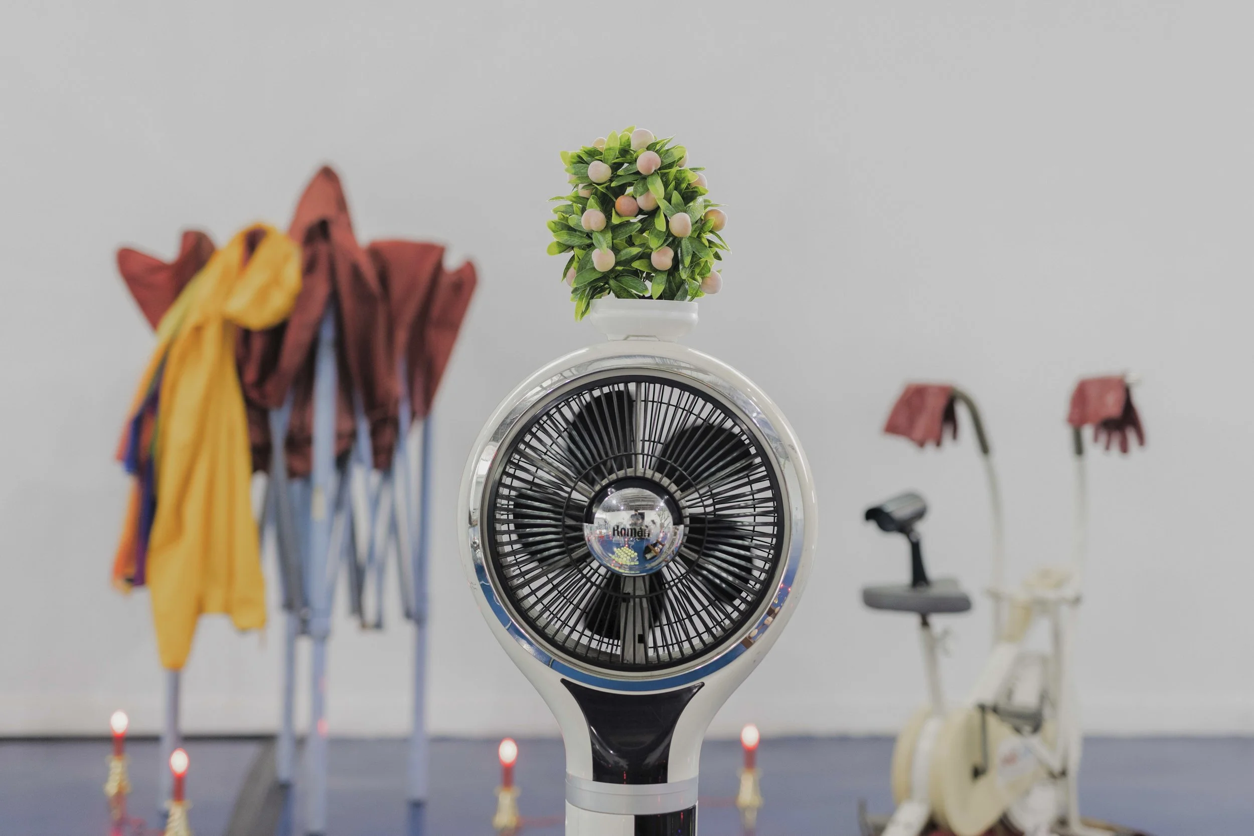 A white fan with a small potted plant on top, in the background are colorful umbrellas, a scooter, and a white bicycle with pink flowers on the handlebar.