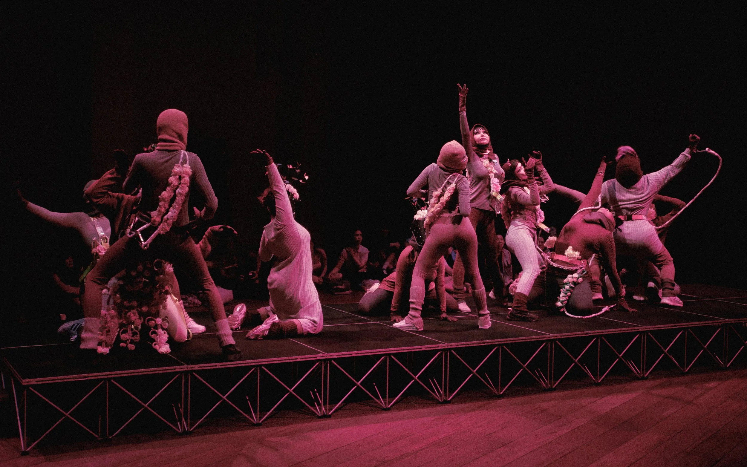 A theatrical stage performance with multiple performers dressed in costumes, some with hoses and floral accessories, on a dark stage with pink lighting.