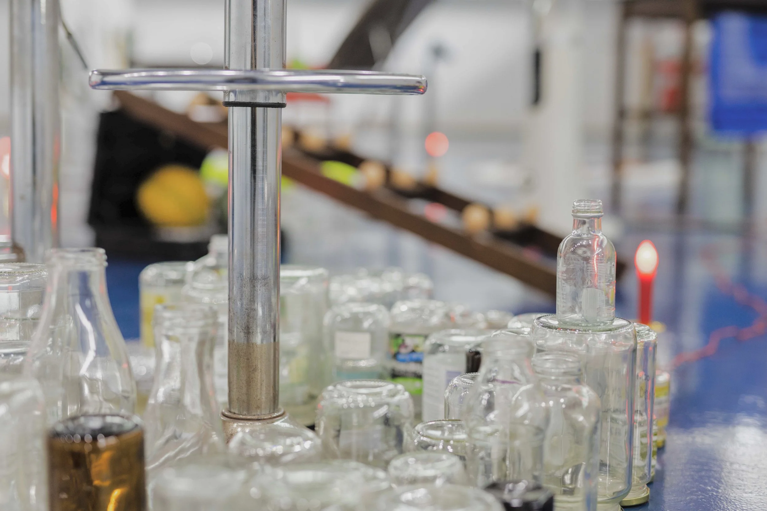 A conveyor belt of glass bottles and jars in a factory or warehouse setting, with a metal pole and a blurred background of a person and other objects.