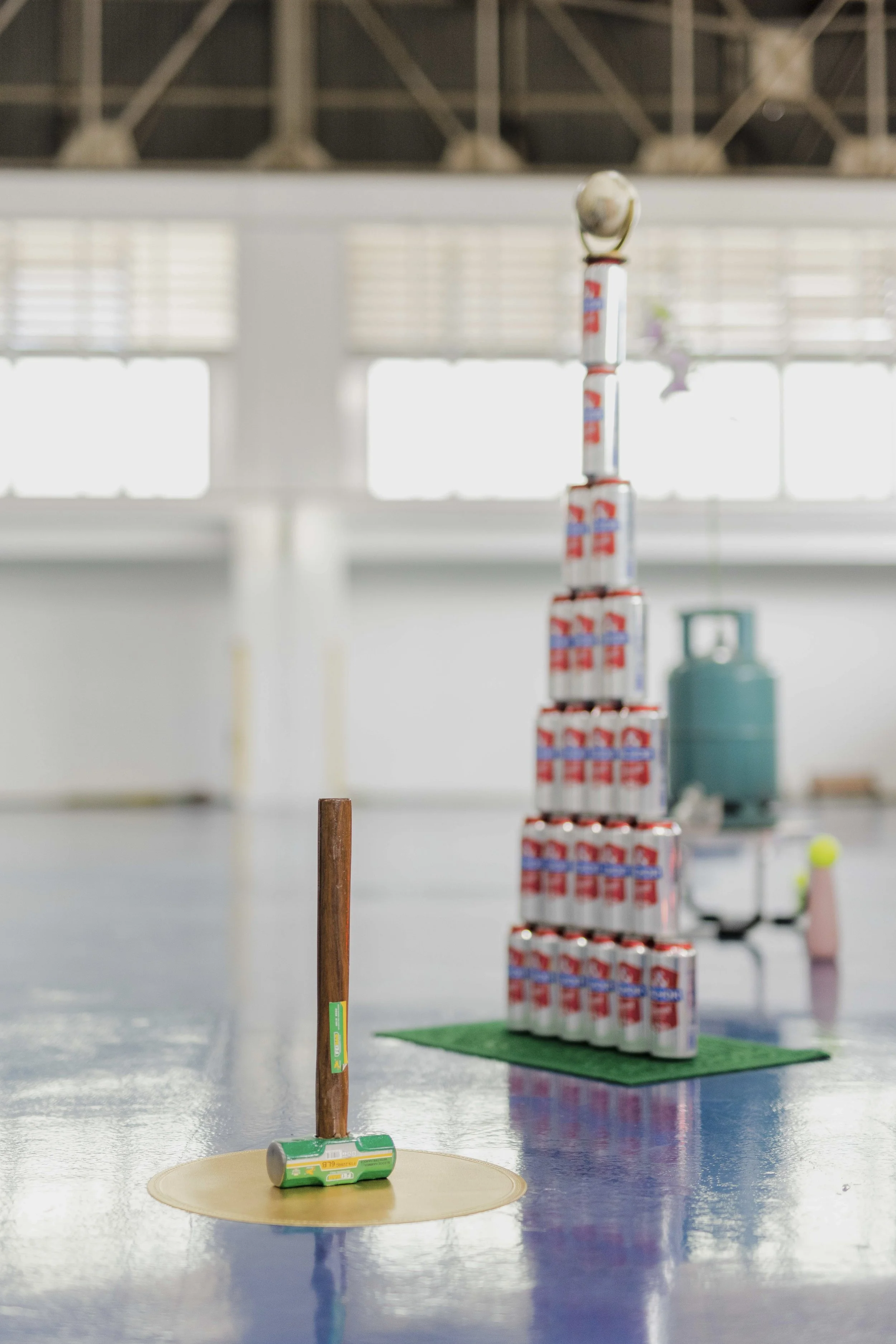 Indoor scene with a stack of soda cans arranged in decreasing size from bottom to top, resembling a tall tower, and a small green and silver toy hammer placed on the floor in front of the tower.