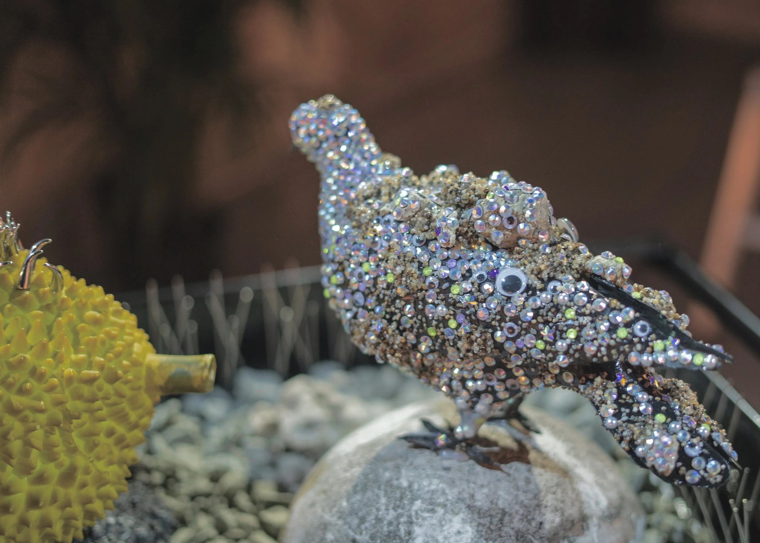 A chameleon sculpture decorated with multicolored rhinestones, perched on a rock, with a yellow plant in the foreground.