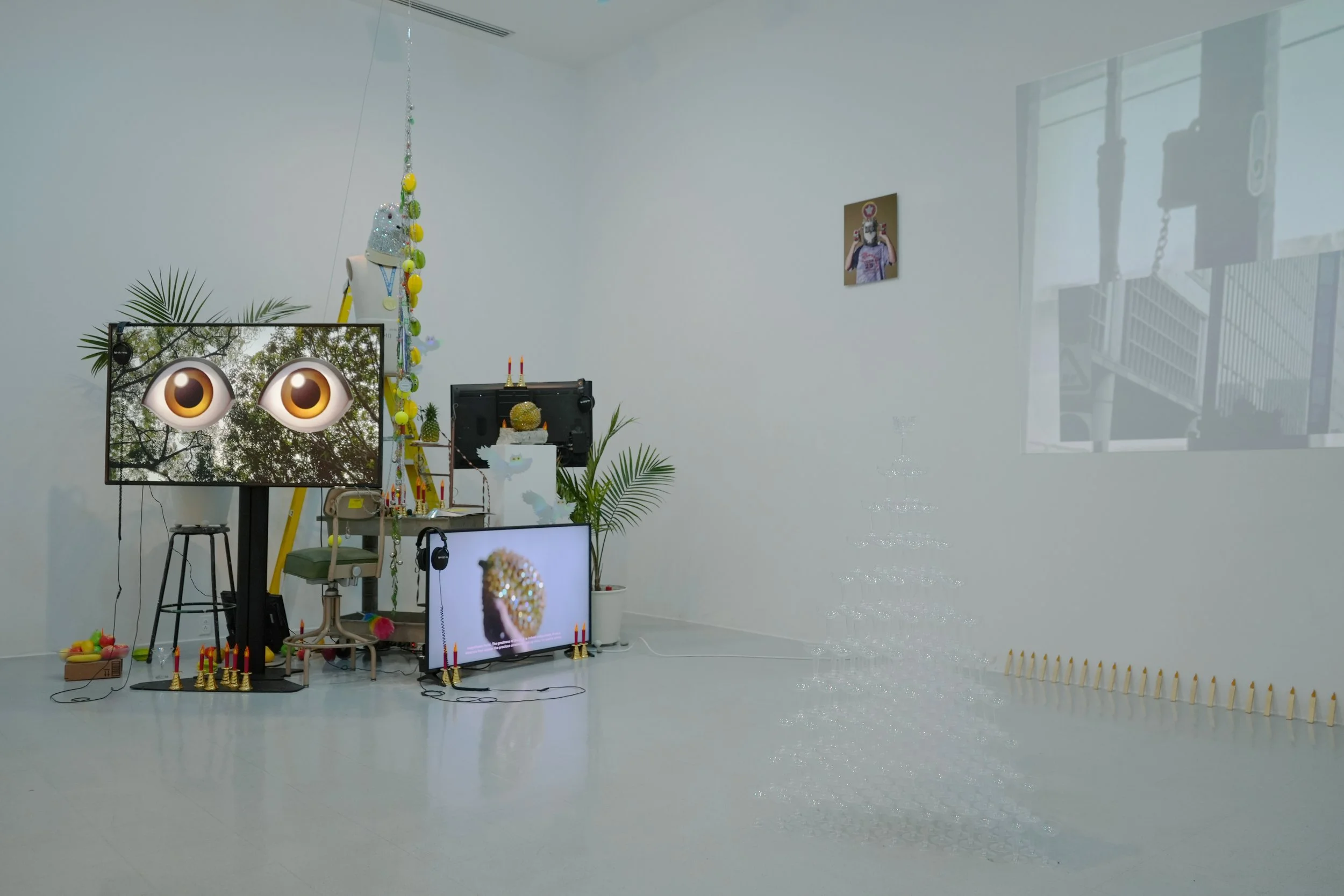 Contemporary art installation in a gallery featuring screens displaying animated eyes and jewelry, adorned with decorative objects, plants, and small candles, with a white wall and window in the background.
