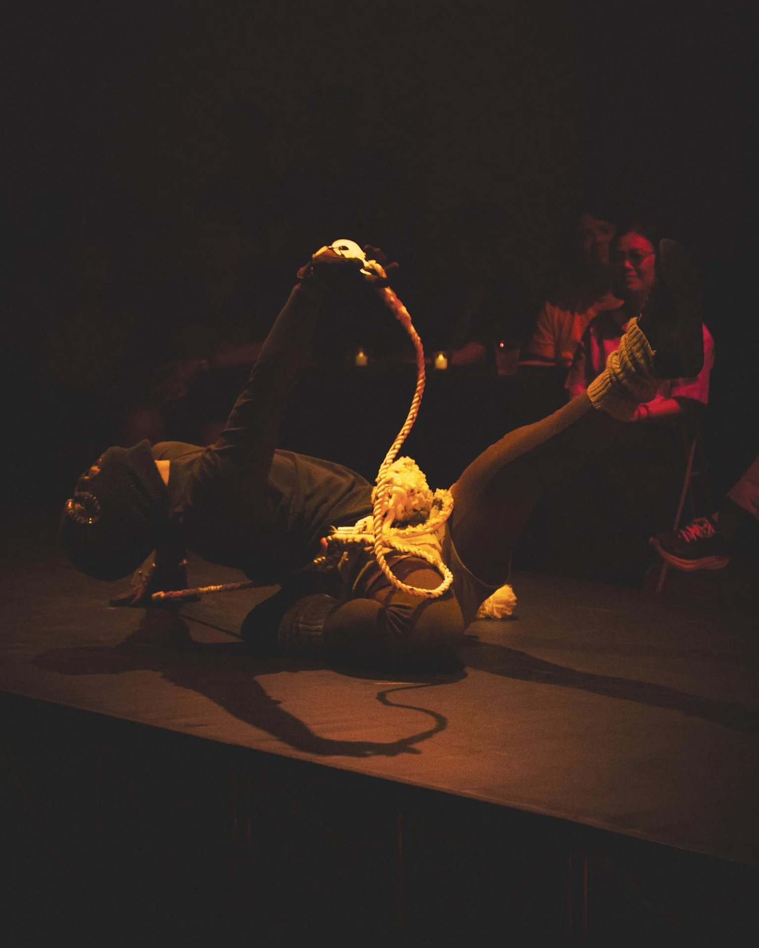 A performer on stage, dressed in black and wearing a mask, is lying on their side with one leg lifted and extended, holding a rope with a circular object at the end. There are people sitting in the background watching the performance in a dimly lit e
