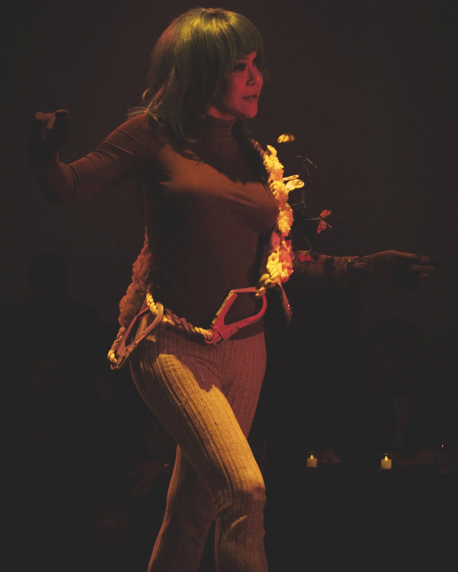 A woman dancing on stage with a relaxed pose, wearing a long-sleeve top, striped pants, and a lei around her neck, illuminated by warm, subdued stage lighting.