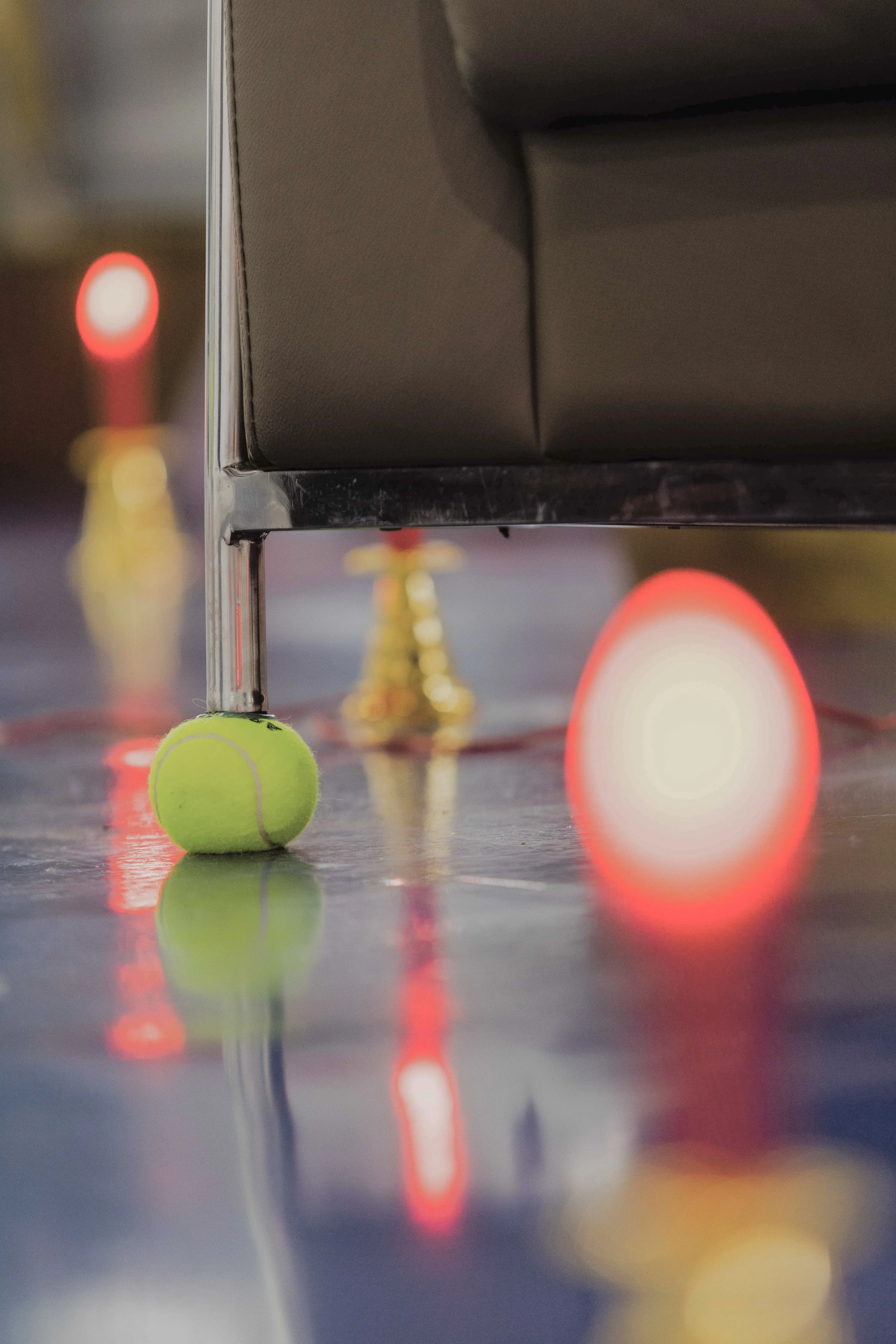 Close-up of a tennis ball under a piece of furniture, with blurred red and yellow lights reflected on the shiny floor.