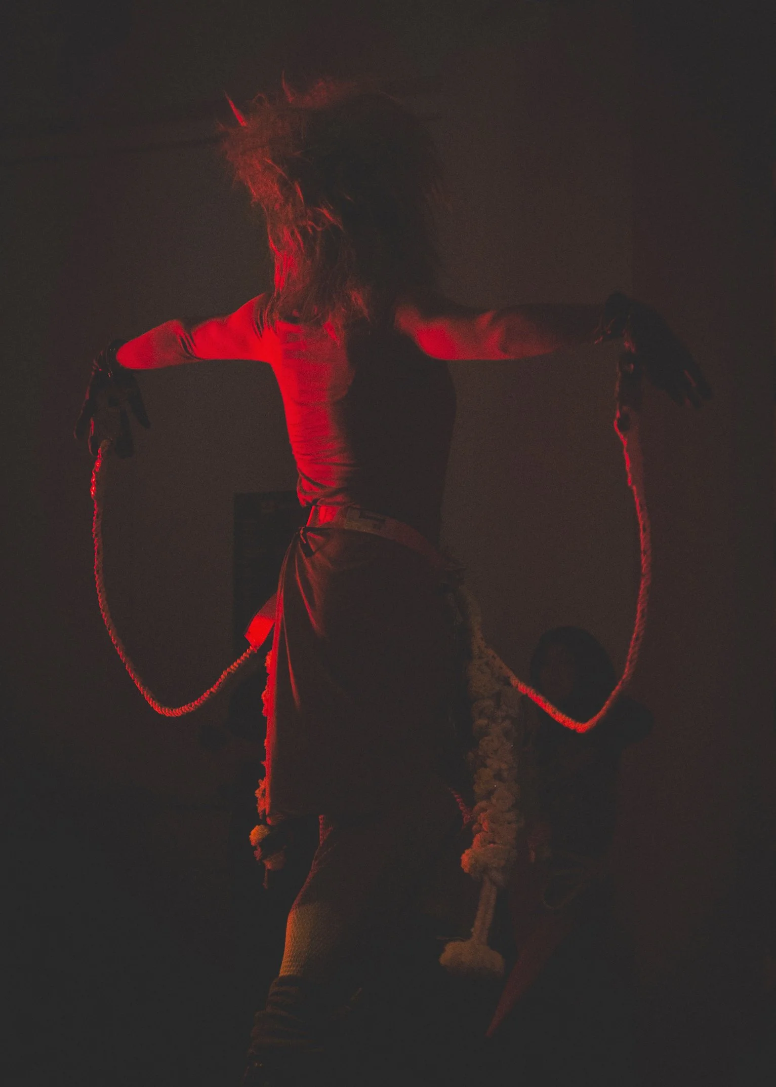 A person with long, wild hair stands with arms outstretched, holding chains in both hands, illuminated by red light in a dark room.