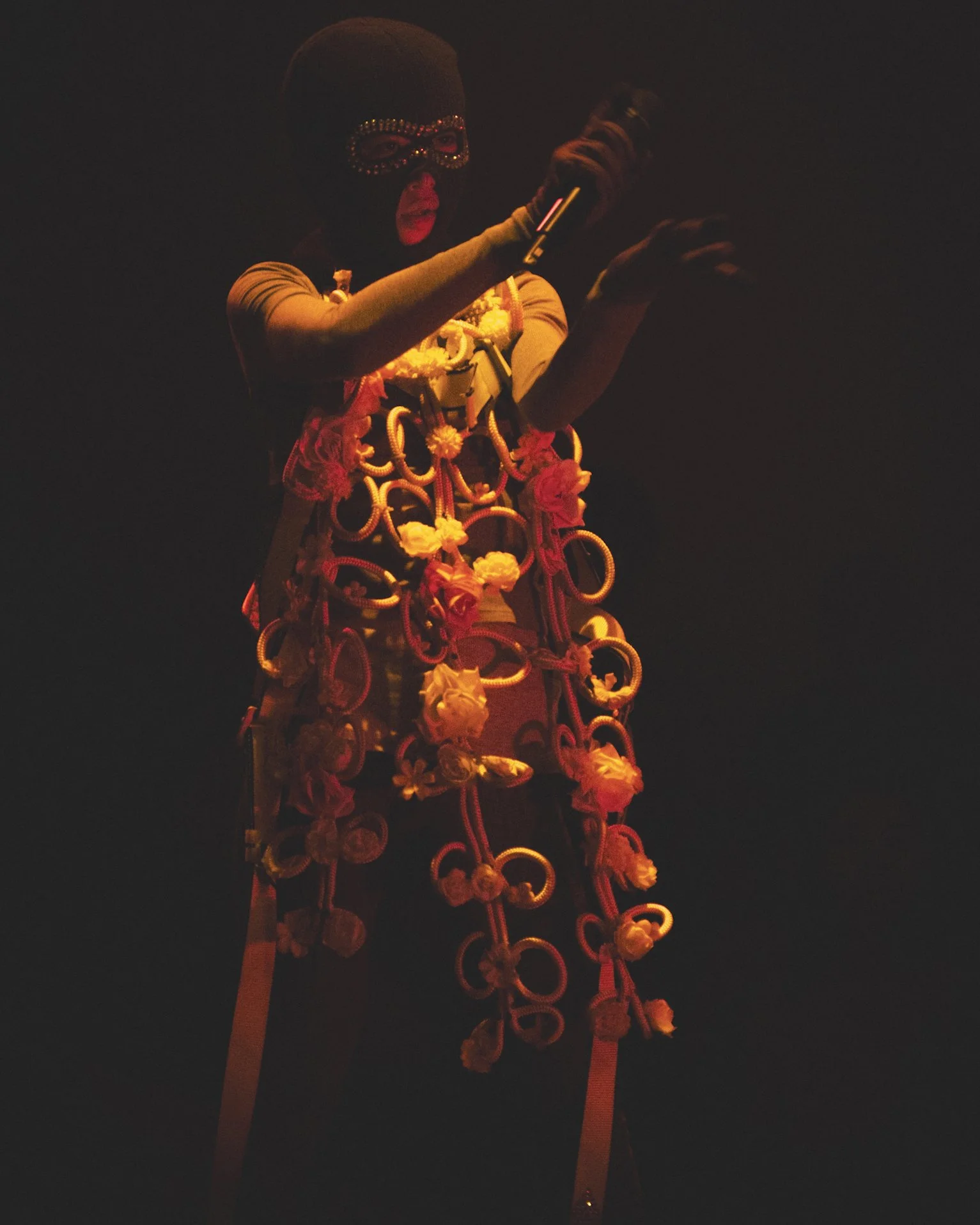 Person wearing a mask and decorated with colorful flowers and rings, holding a microphone, on a stage in a dark setting.