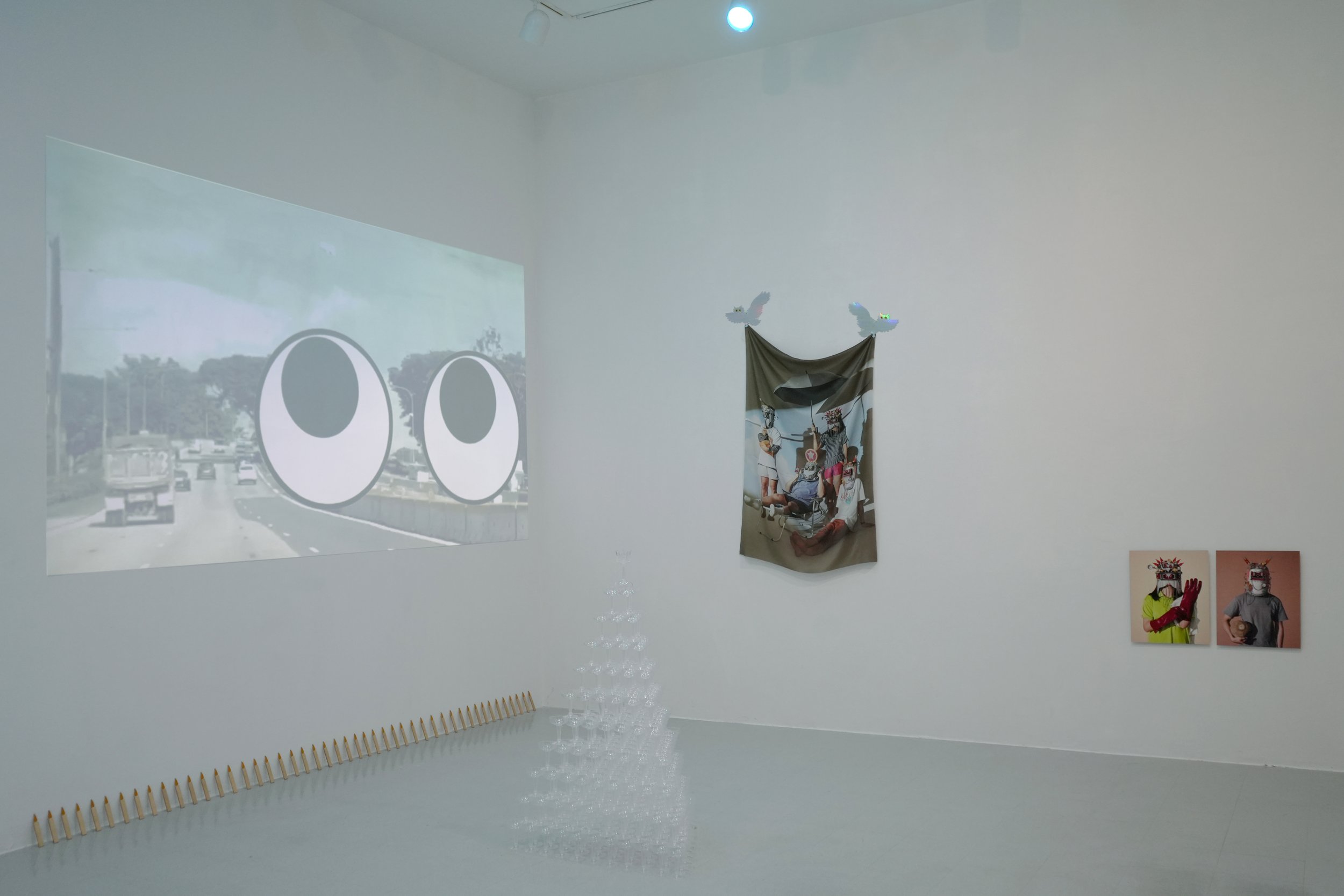 An art installation in a white-walled room featuring a video projection of a busy road with cartoonish eyes, a fabric with a photo of a group of people wearing masks hanging from the ceiling, two small photographs of individuals wearing masks on the 