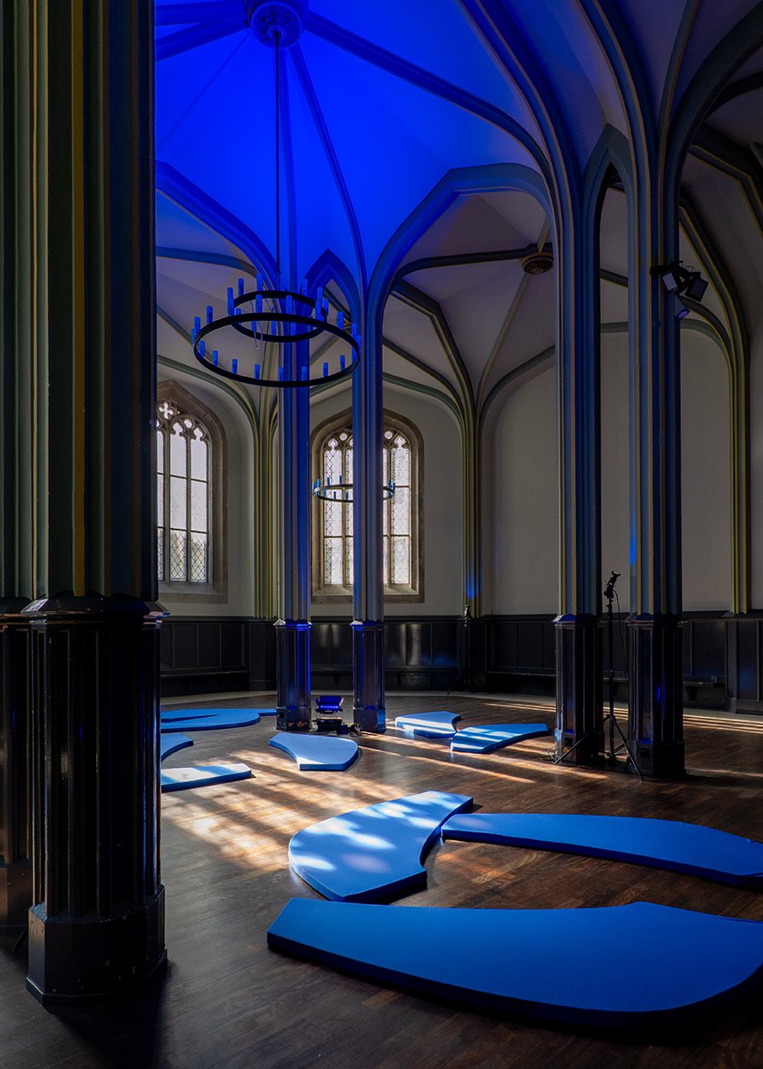 Empty Gothic-style room with tall arched windows, dark wood paneling, and modern blue and black circular chandeliers; blue seating cushions arranged on the floor, with sunlight casting patterned shadows.