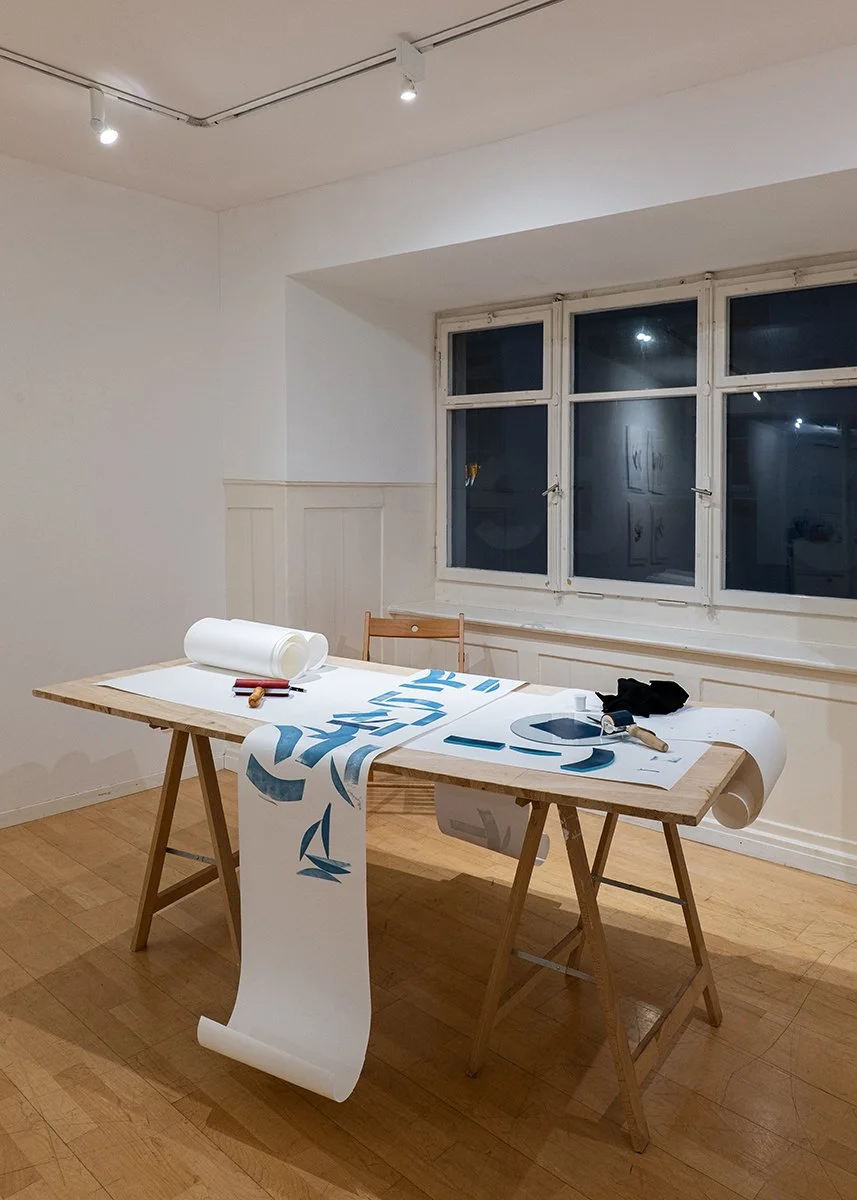A wooden table with large sheets of white paper, some rolled up and some partially unrolled, with blue shapes on them. There are pens, a roller, and a black cloth on the table. The room has white walls, it is a setup for linoleum printing workshop. 