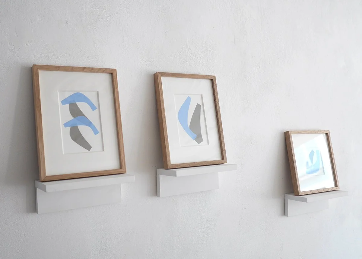 Three framed abstract paper collage art pieces with blue and gray shapes, displayed on white shelves against a white wall.
