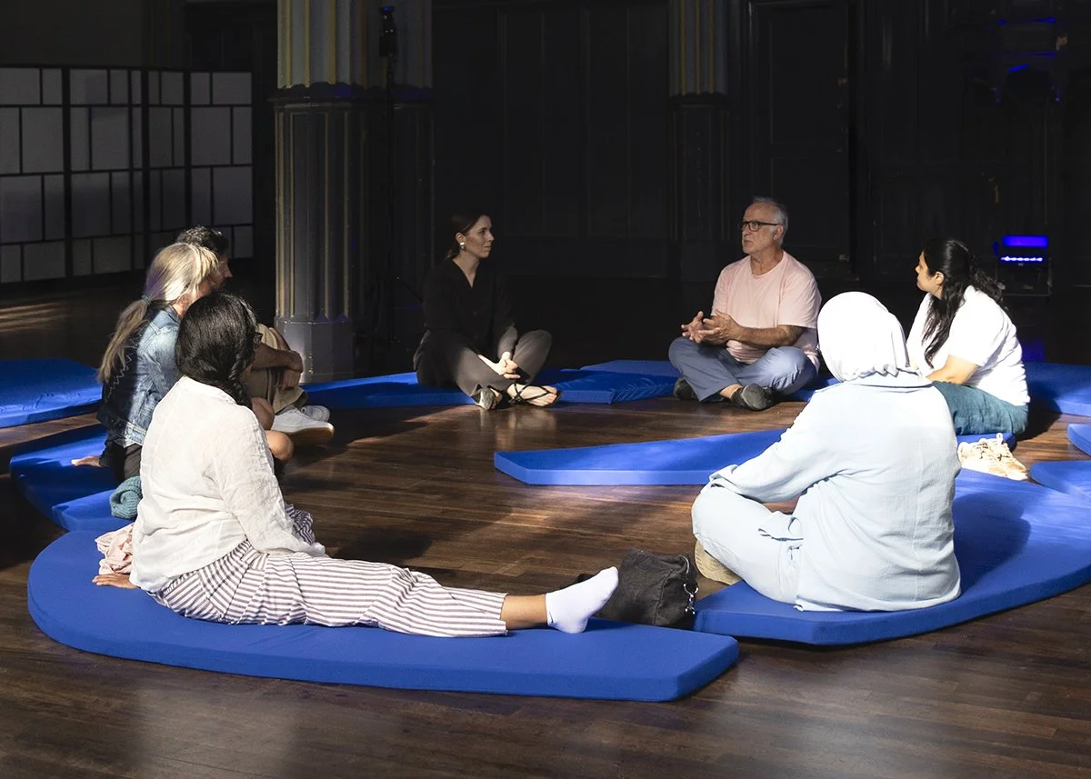 Group of people sitting in a circle on blue cushions in a dark room, engaged in a discussion.