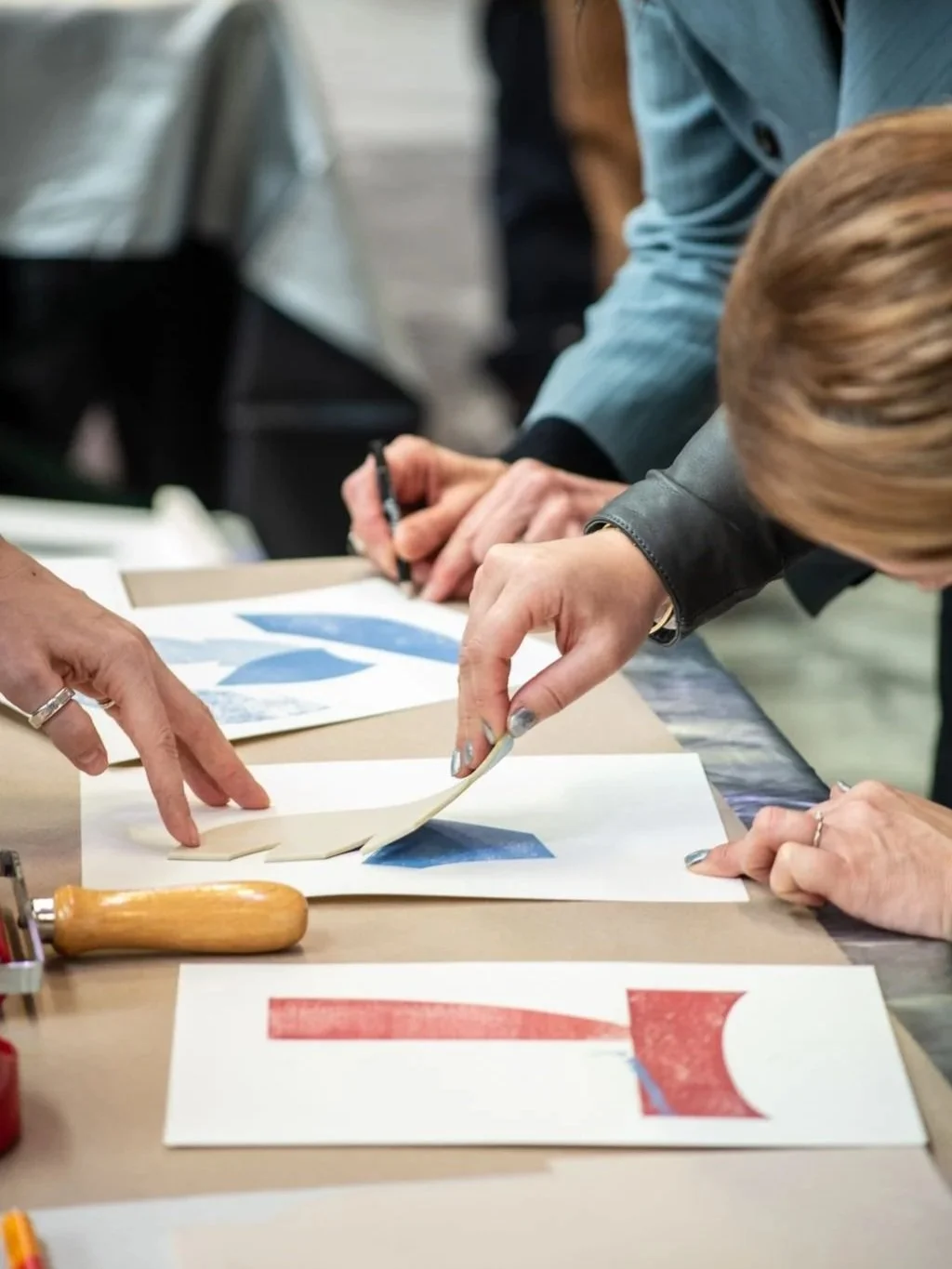 People working on an art project with paper, scissors, and a cutting tool on a table.