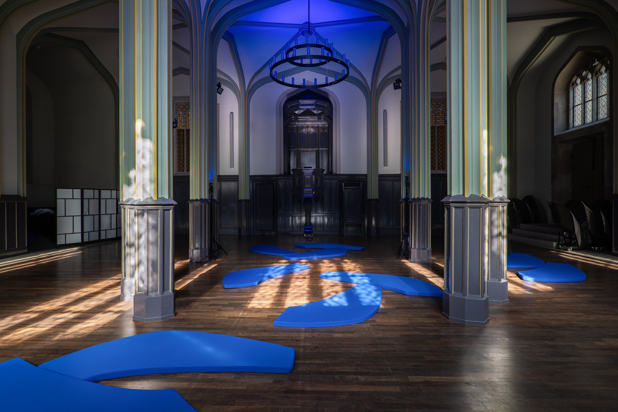 An indoor space with Gothic-style arches, large windows, and wooden floors, featuring blue foam shapes arranged on the floor and a dark wooden altar or stage at the back.