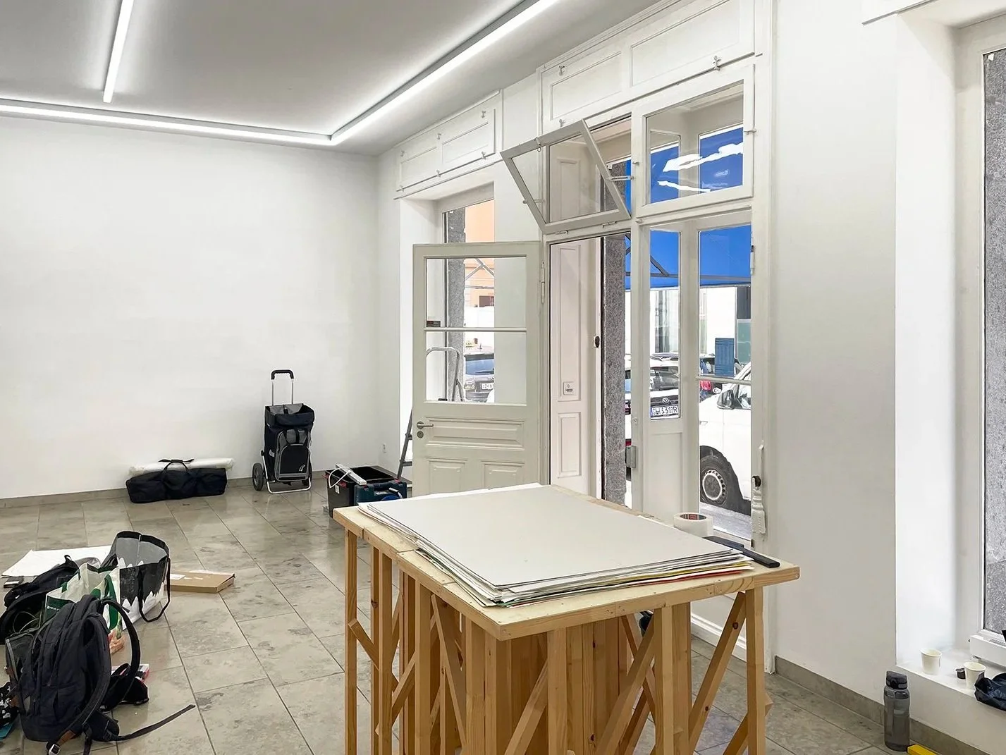 Empty store or office space with a wooden table holding large sheets of paper, open door and windows, rolling cart, bags, and construction tools on the tiled floor.