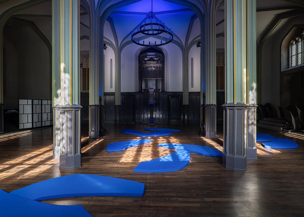 Interior of a room with tall, arched ceilings, illuminated by natural light through windows, and featuring modern blue floor decorations resembling abstract shapes or paths on a wooden floor.